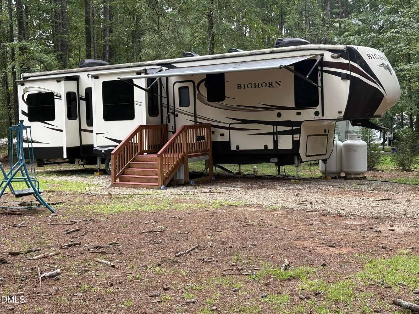 A large Bighorn fifth-wheel RV with a set of wooden stairs sits parked on a gravel and dirt area surrounded by tall trees in a forested campsite at 103 Creek Drive, Louisburg.