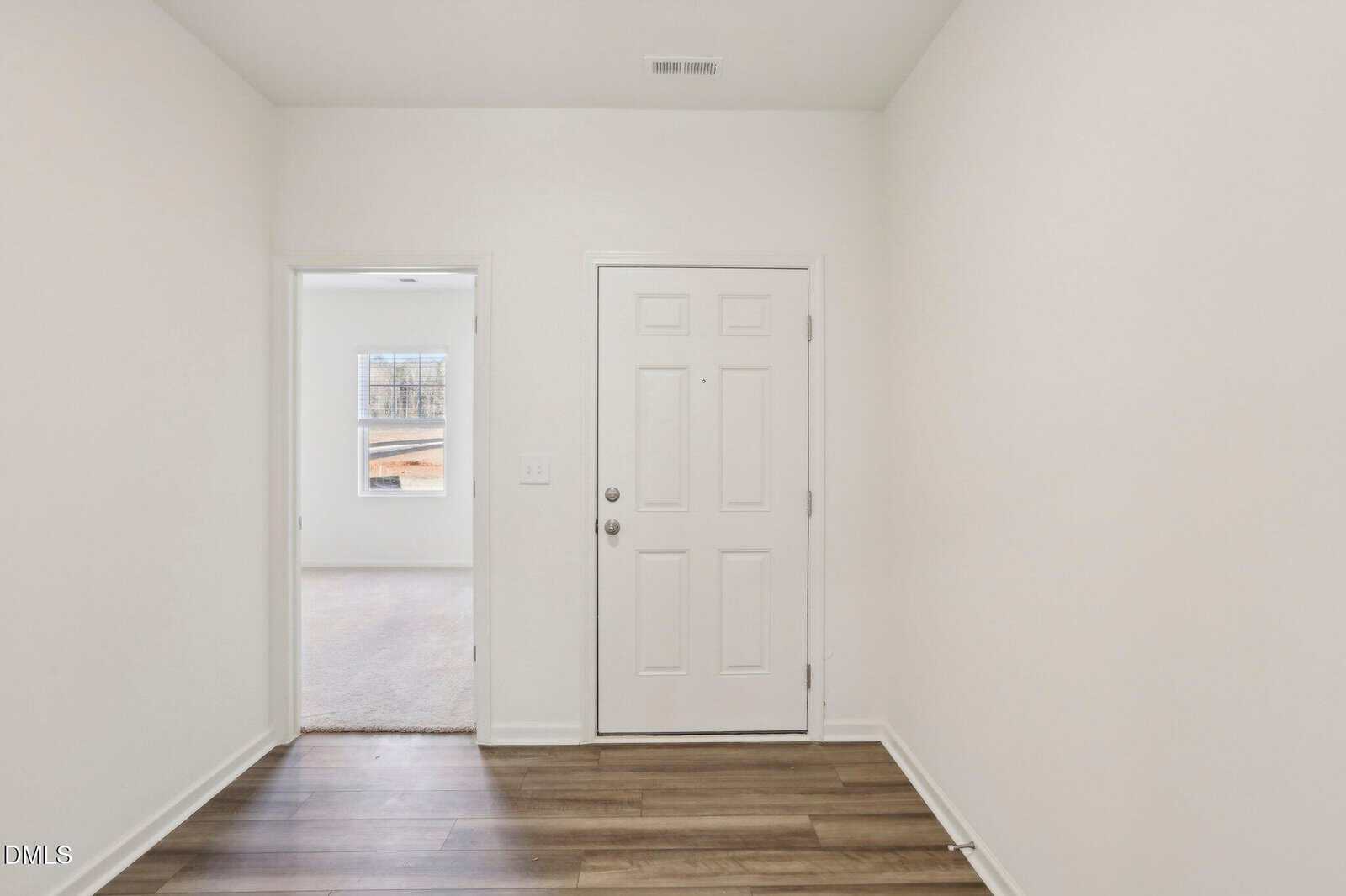 A white front door with a peephole and two locks is centered on a plain wall. Brown wood-style flooring leads to a doorway on the left, revealing a carpeted room with a window. Walls and ceiling are white.