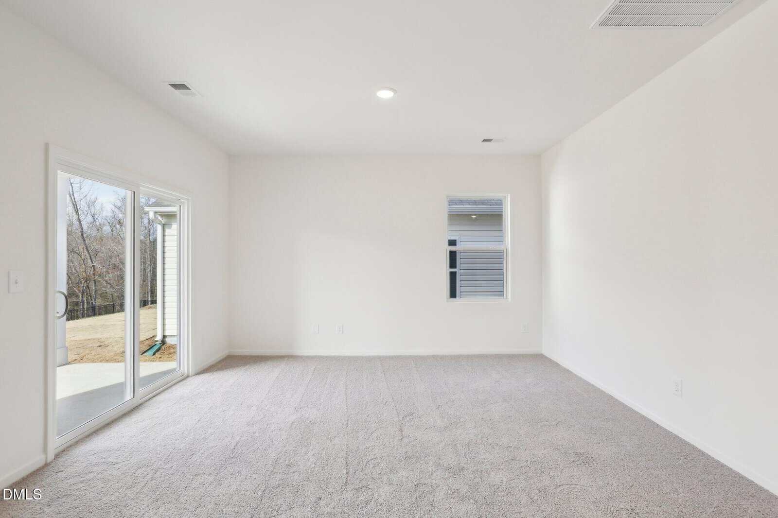 A room with light-colored carpet, white walls, a sliding glass door leading outside, and a window on the right wall. The space is empty and well-lit by natural light.
