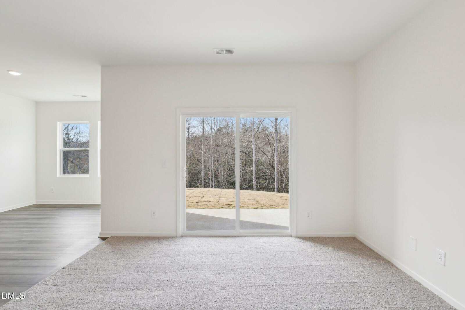 A bright, empty room with white walls and carpet, featuring a large sliding glass door opening to a patio and a view of trees. Adjacent space has wood flooring and a window.