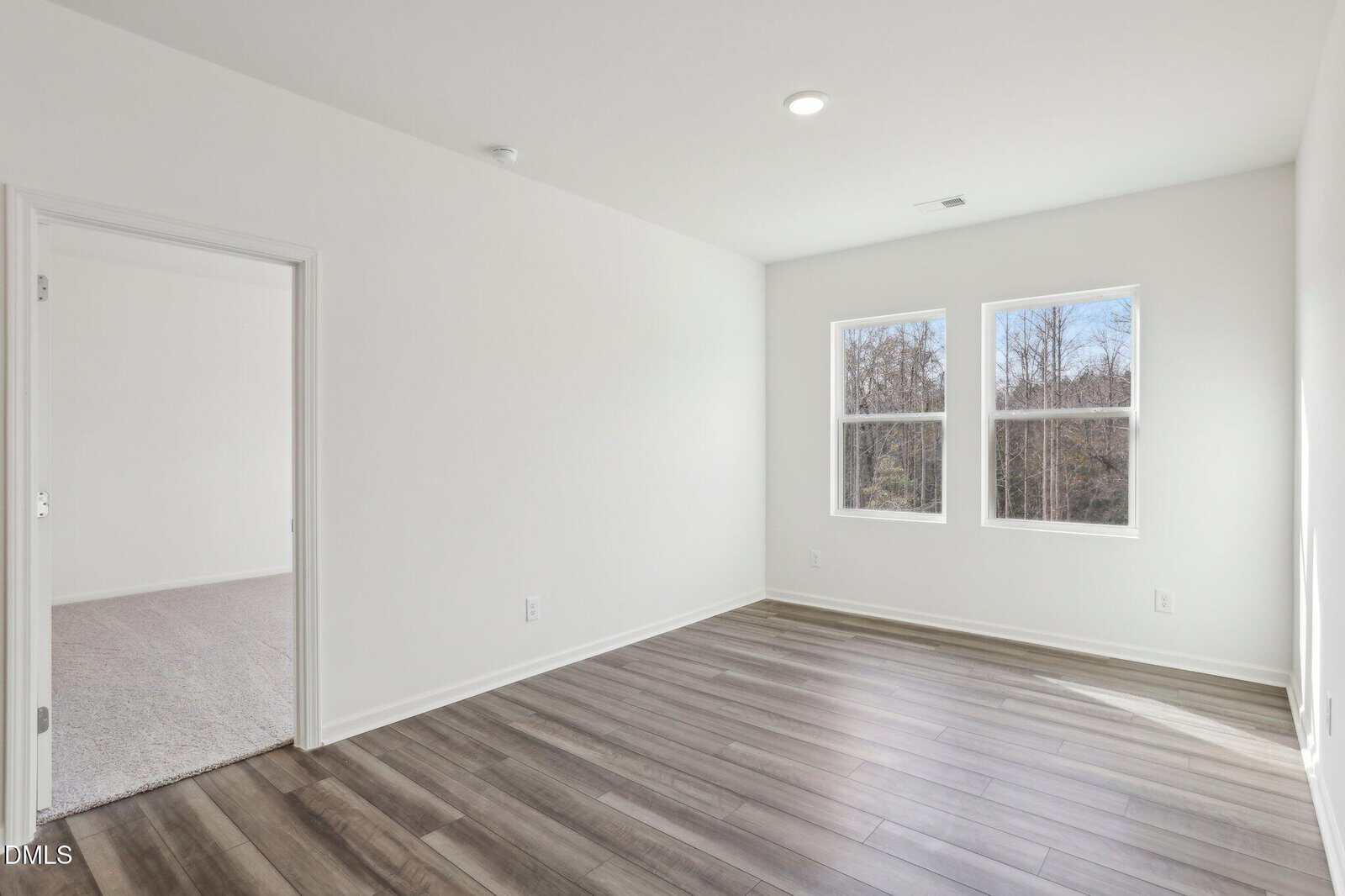 Empty, unfurnished room with white walls, wood-style flooring, two large windows letting in natural light, and an open doorway leading to a carpeted area.