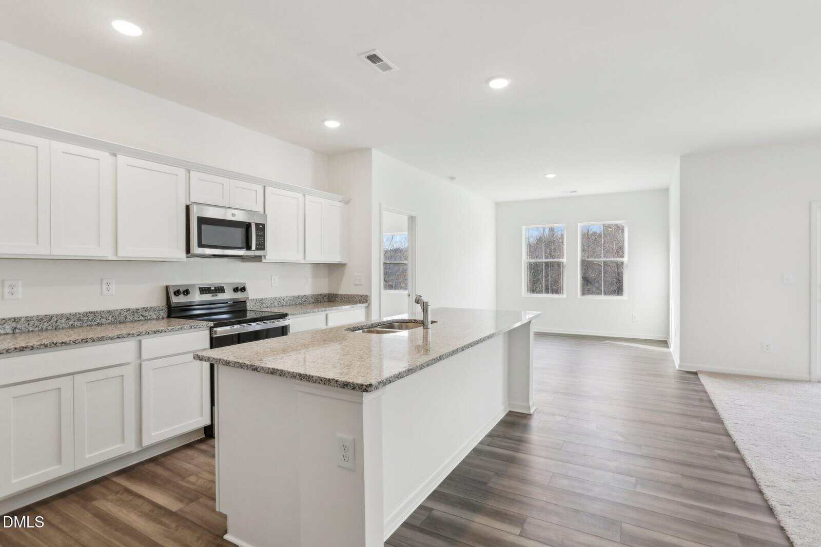 Modern kitchen with white cabinets, stainless steel appliances, a large granite island, and open layout leading to a bright living area with large windows and wood-style flooring.