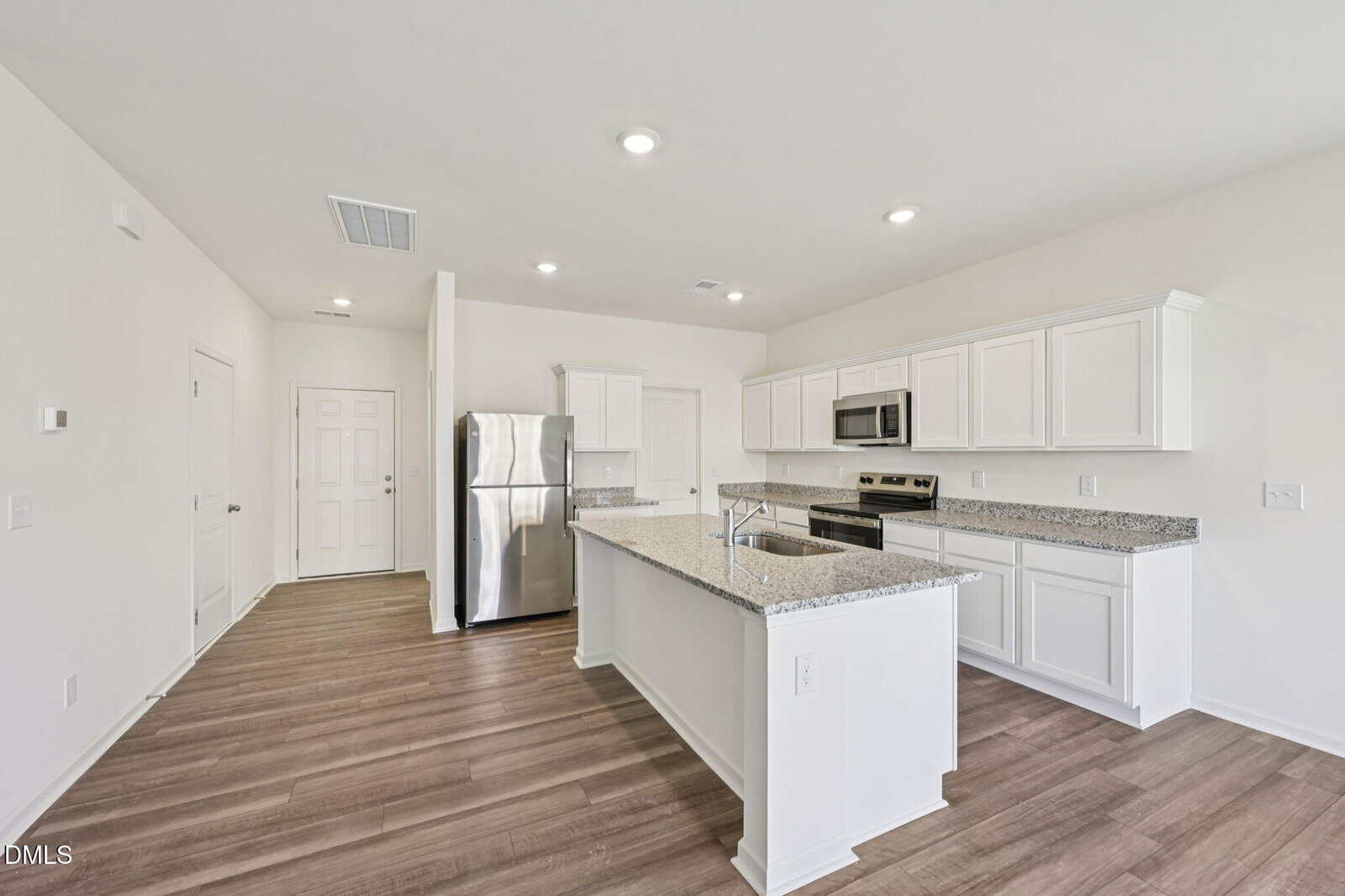 Modern kitchen with white cabinets, stainless steel appliances, a central island with a sink, and light wood flooring. The walls and ceiling are painted white, and recessed ceiling lights provide illumination. Door and entryway visible in background.