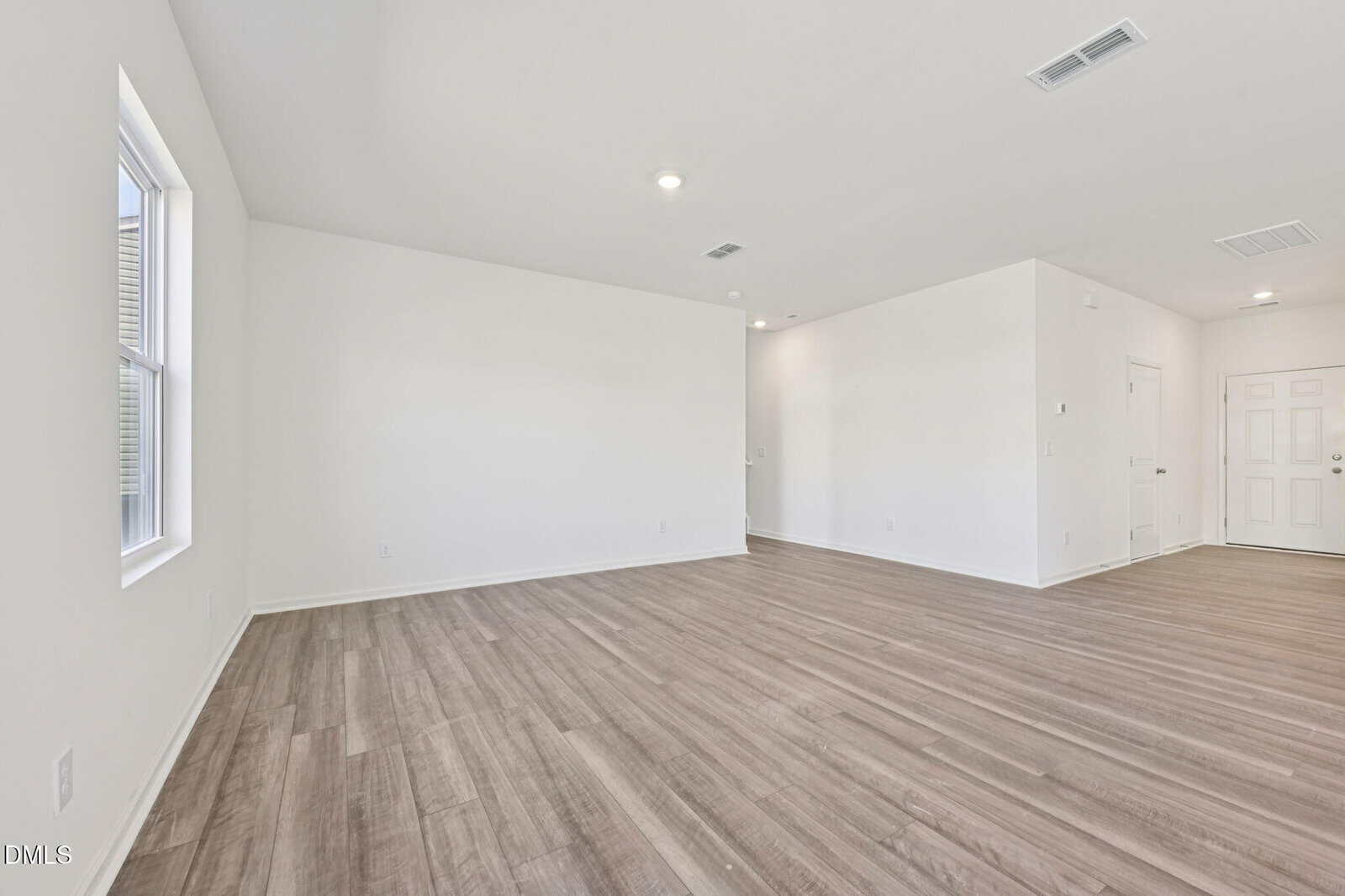 Unfurnished, empty room with light wood flooring, white walls, and ceiling lights. A window is on the left wall, and a white entry door is in the background. The space is open and modern.