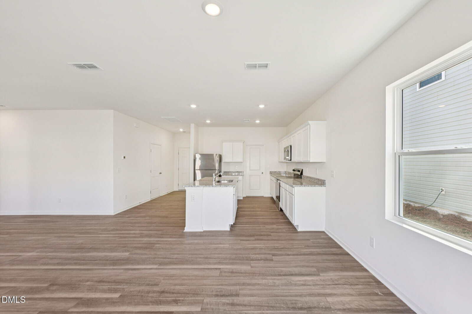 Open-concept kitchen with white cabinets, stainless steel appliances, and granite countertops. The room features wood-style flooring, white walls, recessed lighting, and a large window letting in natural light.