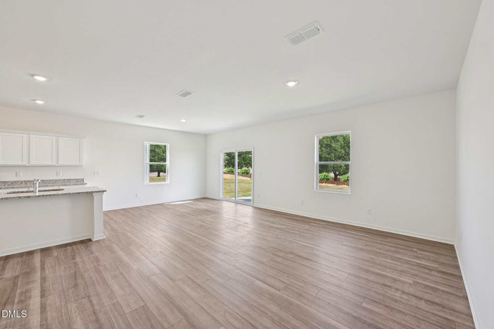 Spacious empty room with light wood flooring, white walls, recessed ceiling lights, two windows, glass doors to the outside, and a kitchen area with white cabinets and a granite countertop.