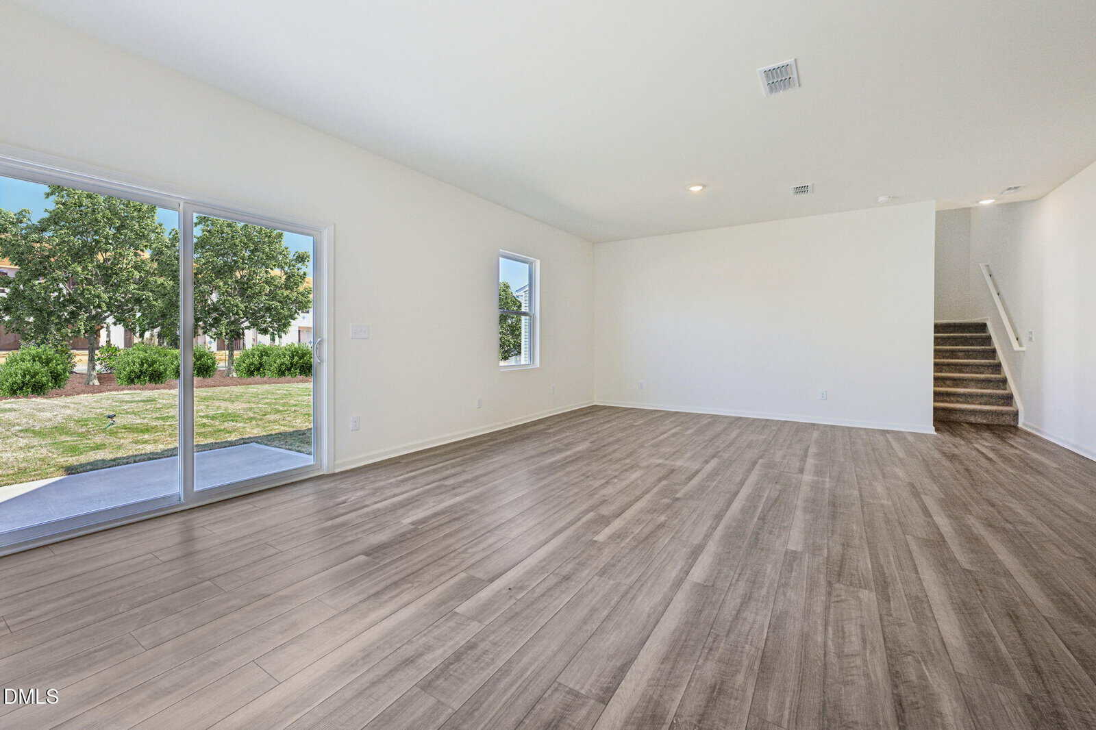 Spacious empty living room with light wood floors, white walls, a small window, sliding glass doors leading to a grassy yard, and carpeted stairs in the back right corner.