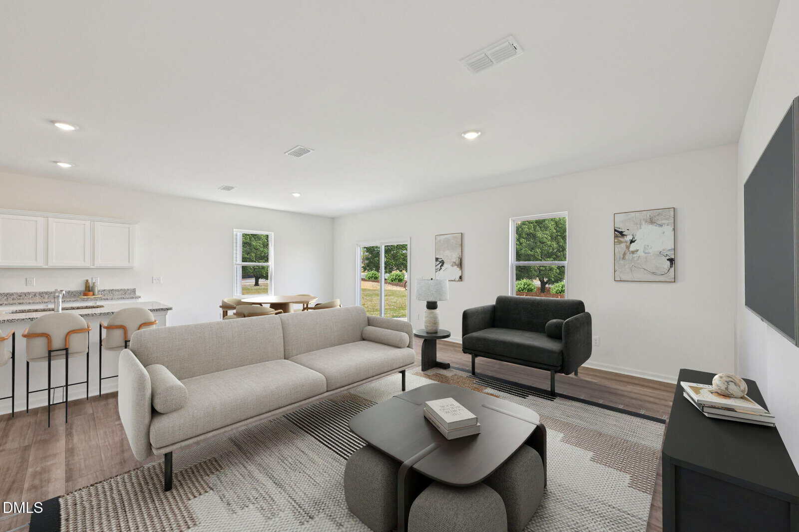 Modern living room with neutral-colored sofas, a black armchair, a coffee table, and wall art. Large windows let in natural light. Open kitchen and dining area with barstools visible in the background.