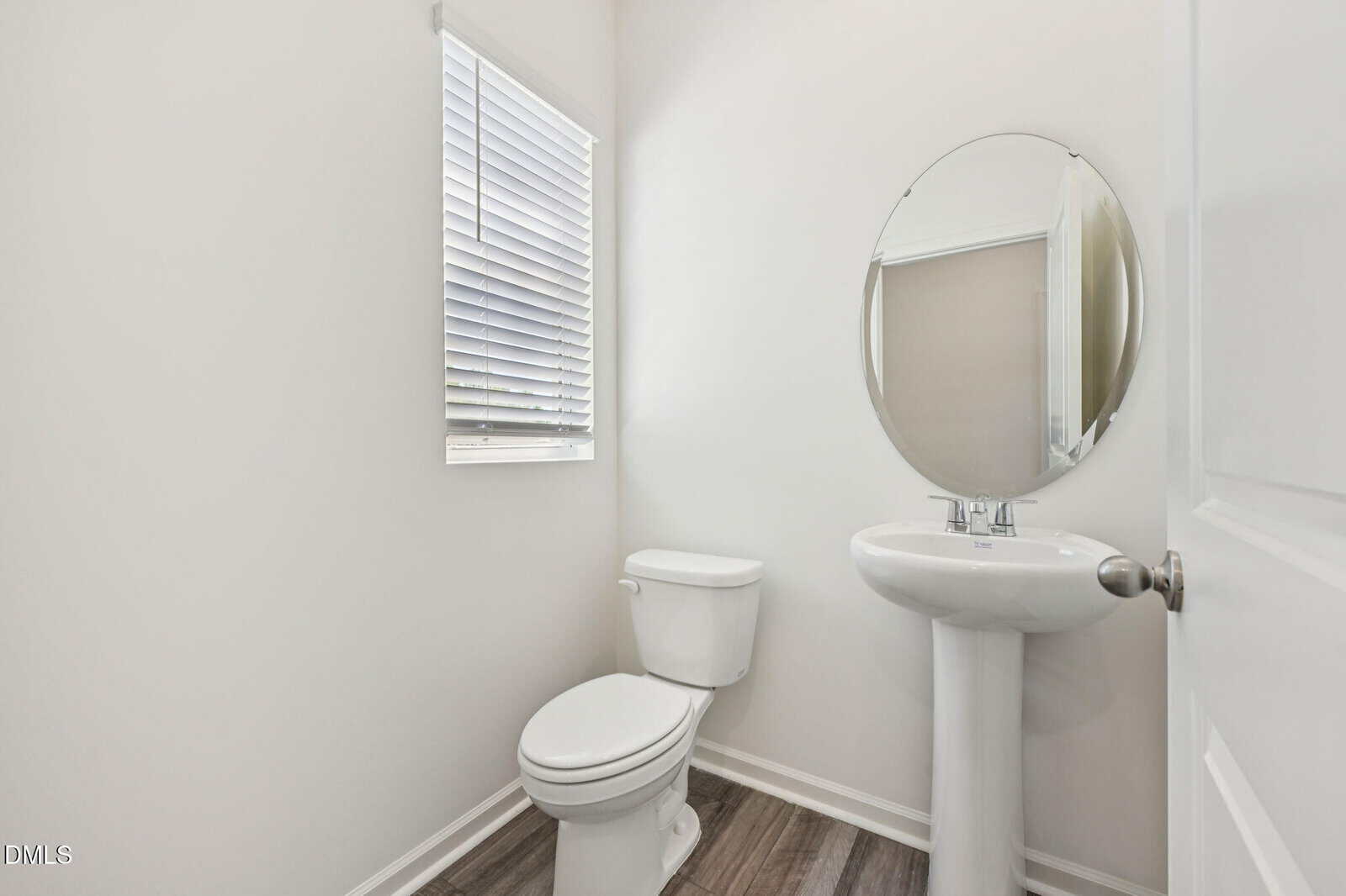 A small bathroom with a white toilet, a pedestal sink with an oval mirror above it, and a window with blinds. The walls are light-colored and the floor has wood-style flooring.