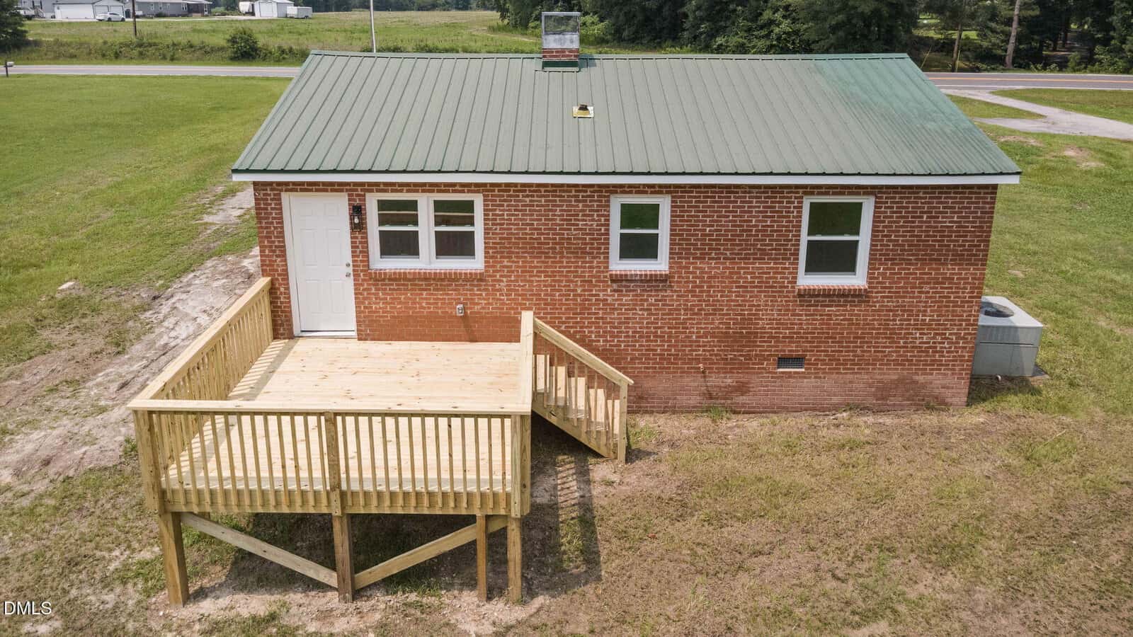 A small brick house with a green metal roof and three windows is shown. A wooden deck with railings extends from the side door, overlooking a grassy yard. A road is visible in the background.