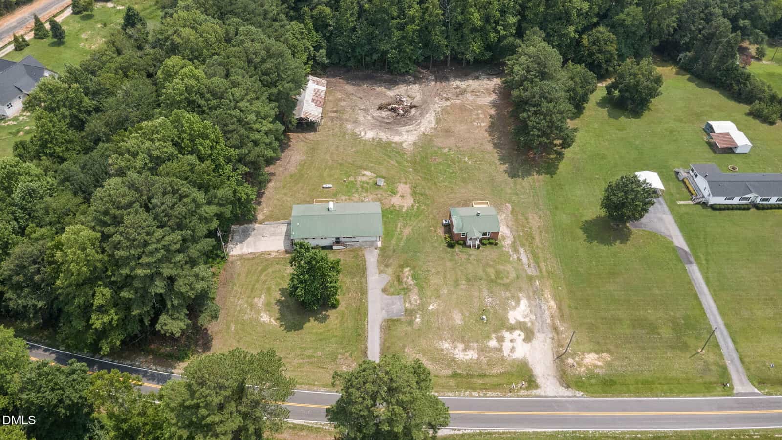 Aerial view of a rural property with two green-roofed buildings, a driveway, open grassy areas, and surrounding trees. The property sits along a curved road, with neighboring homes and additional trees nearby.