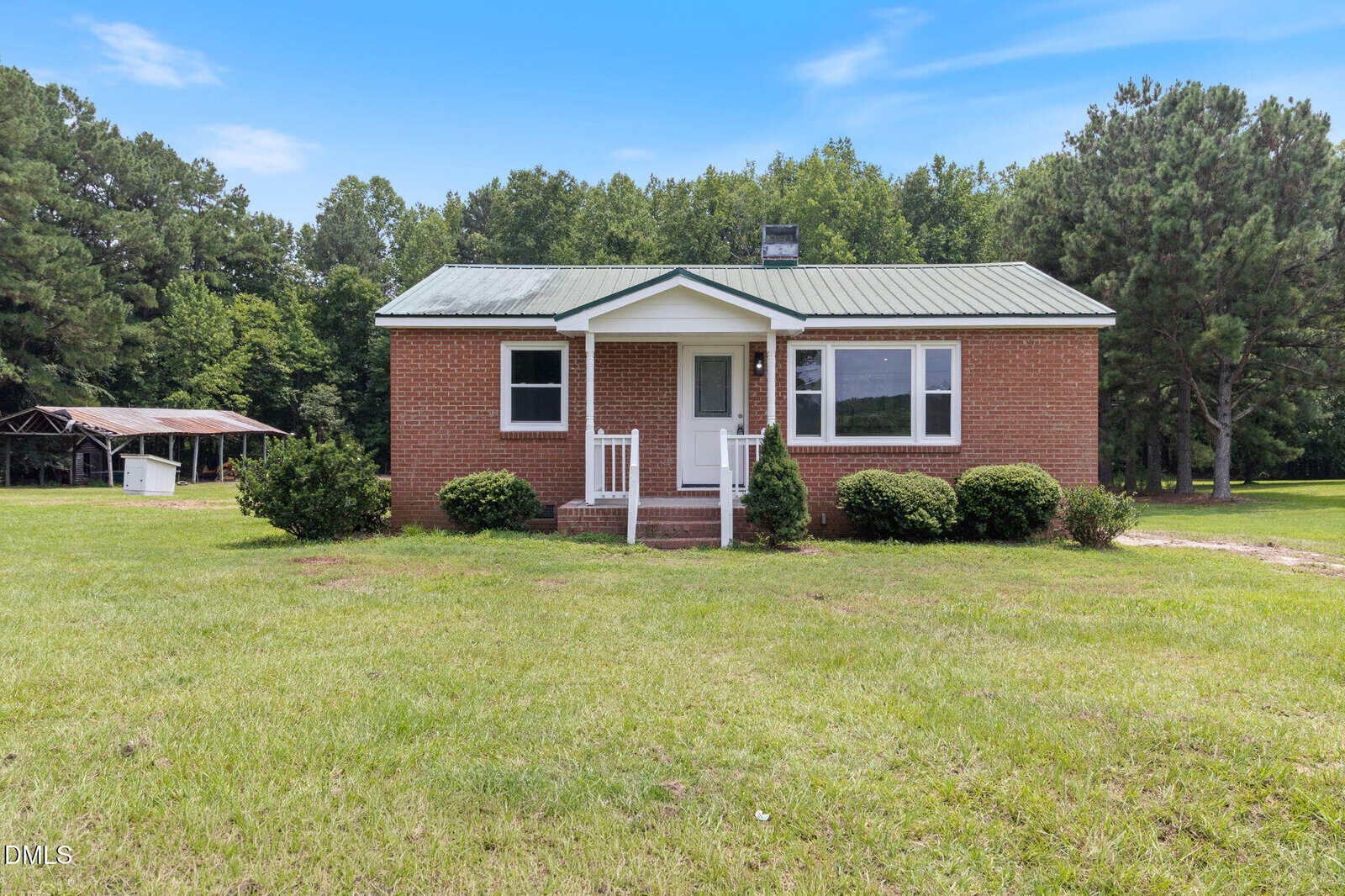 A small brick house with a metal roof sits on a grassy yard at 505 Gay Town Road in Zebulon. There are white railings by the front steps, bushes along the front, and trees in the background. A shed is visible on the left side of the image.