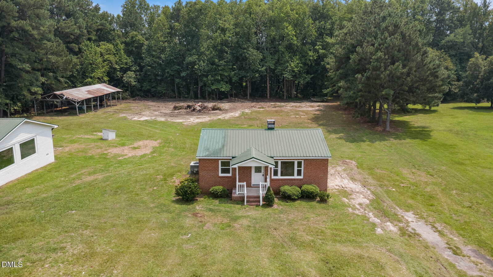 A small brick house with a green metal roof sits in a grassy yard. A dirt driveway leads to the house, and a wooded area is in the background. To the left is a white outbuilding and an open shed structure.