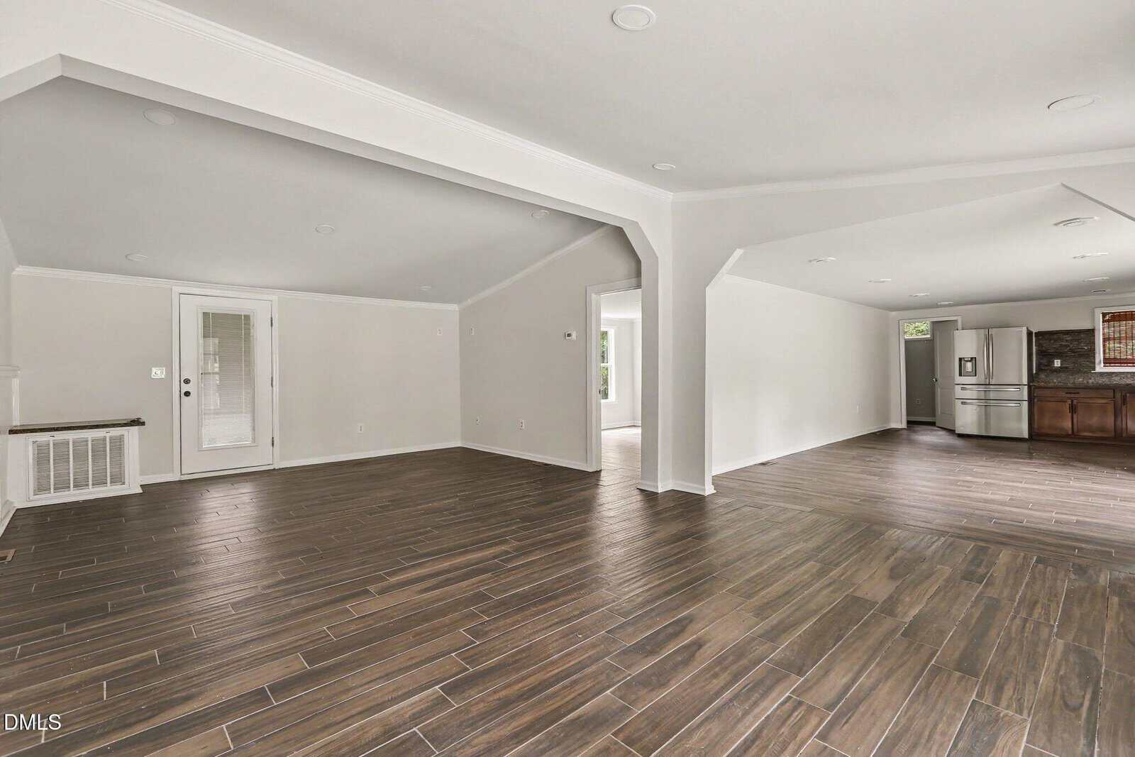Spacious open-concept living area with brown wood-look tile flooring, white walls, recessed lighting, and a view into a kitchen with stainless steel appliances and dark cabinets. Large windows provide natural light.