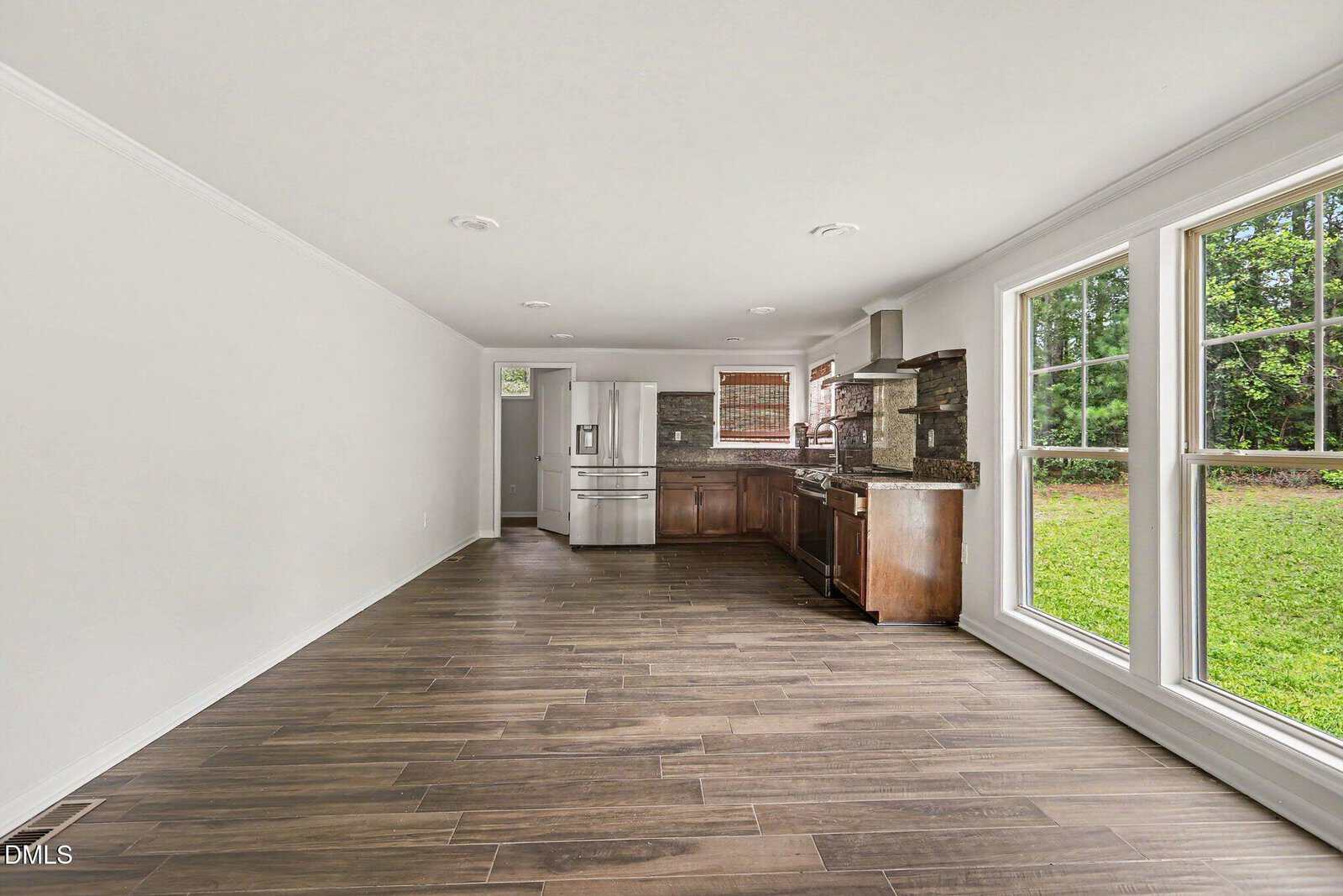 Open kitchen with stainless appliances, dark wood cabinets, and brown tile flooring. Large windows on the right provide a view of green grass and trees outside. White walls and ample natural light fill the space.