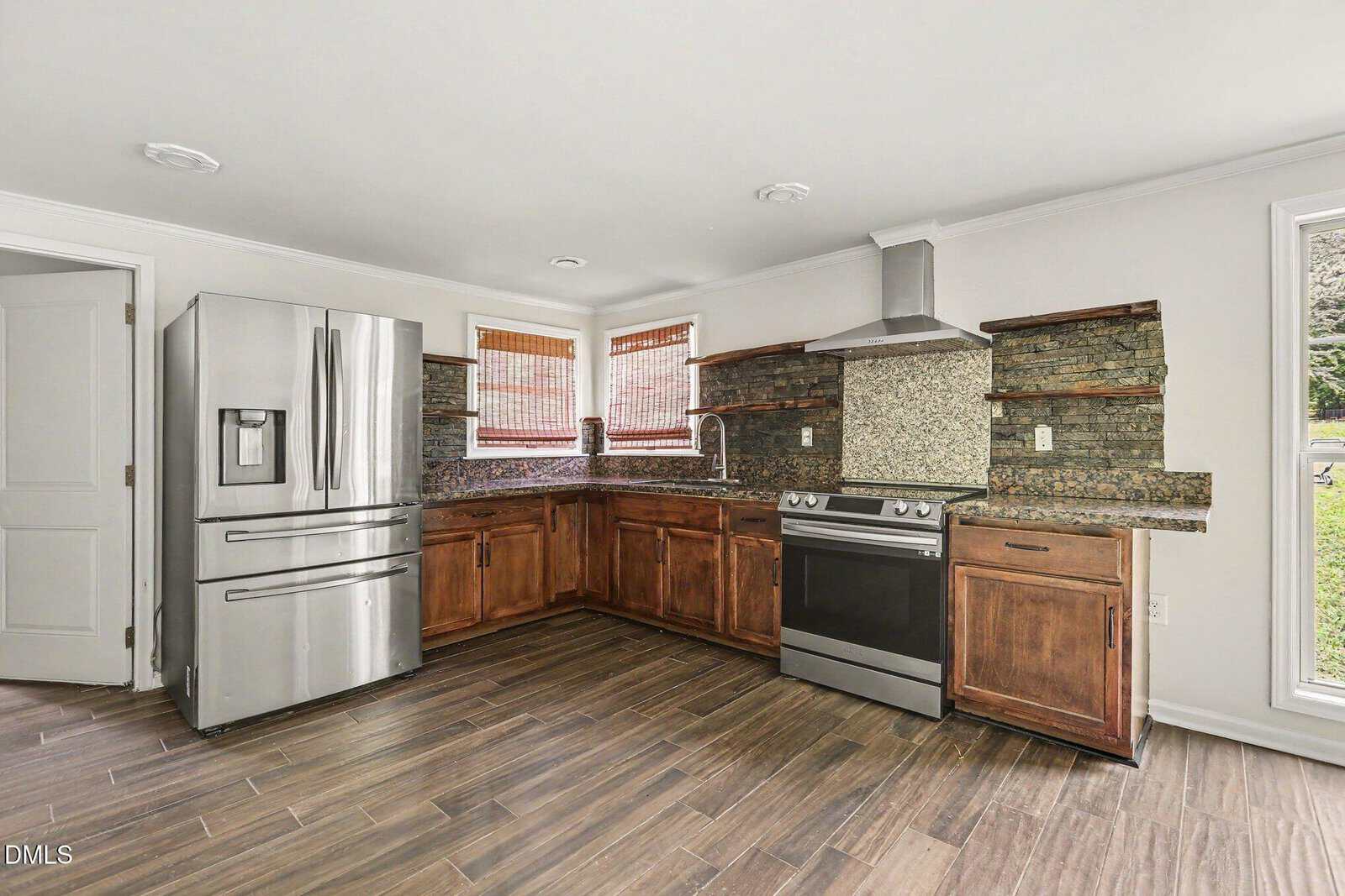 Modern kitchen with stainless steel refrigerator, oven, and range hood. Brown wooden cabinets, mosaic tile backsplash, and two windows with blinds above the sink. The floor is wood-look tile.