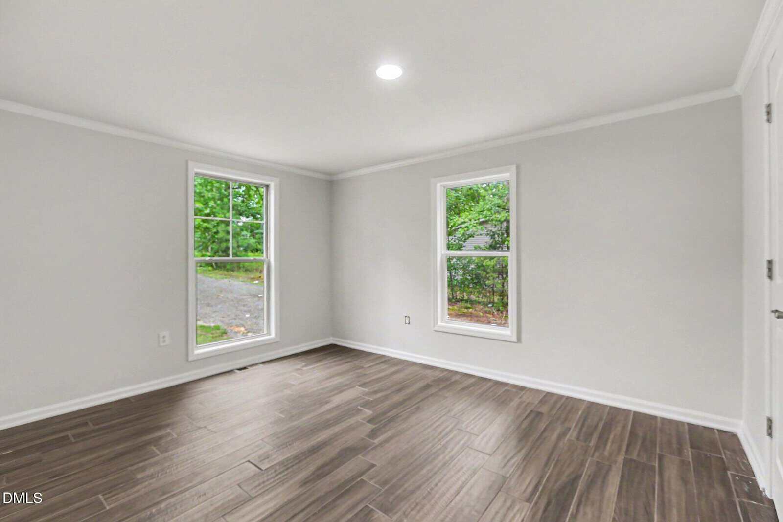 Bright, empty room with light gray walls, wood-look tile flooring, two large windows, white trim, and a ceiling light. Trees are visible through the windows.