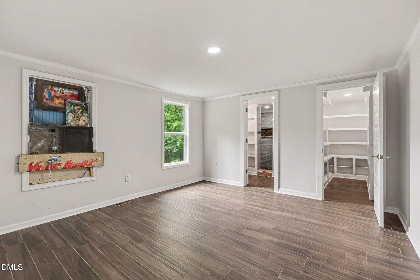 Bright room with wood-look tile flooring, white walls, a window with a green view, an open closet with built-in shelves, and a colorful boarded window on one wall. Ceiling light fixture illuminates the space.
