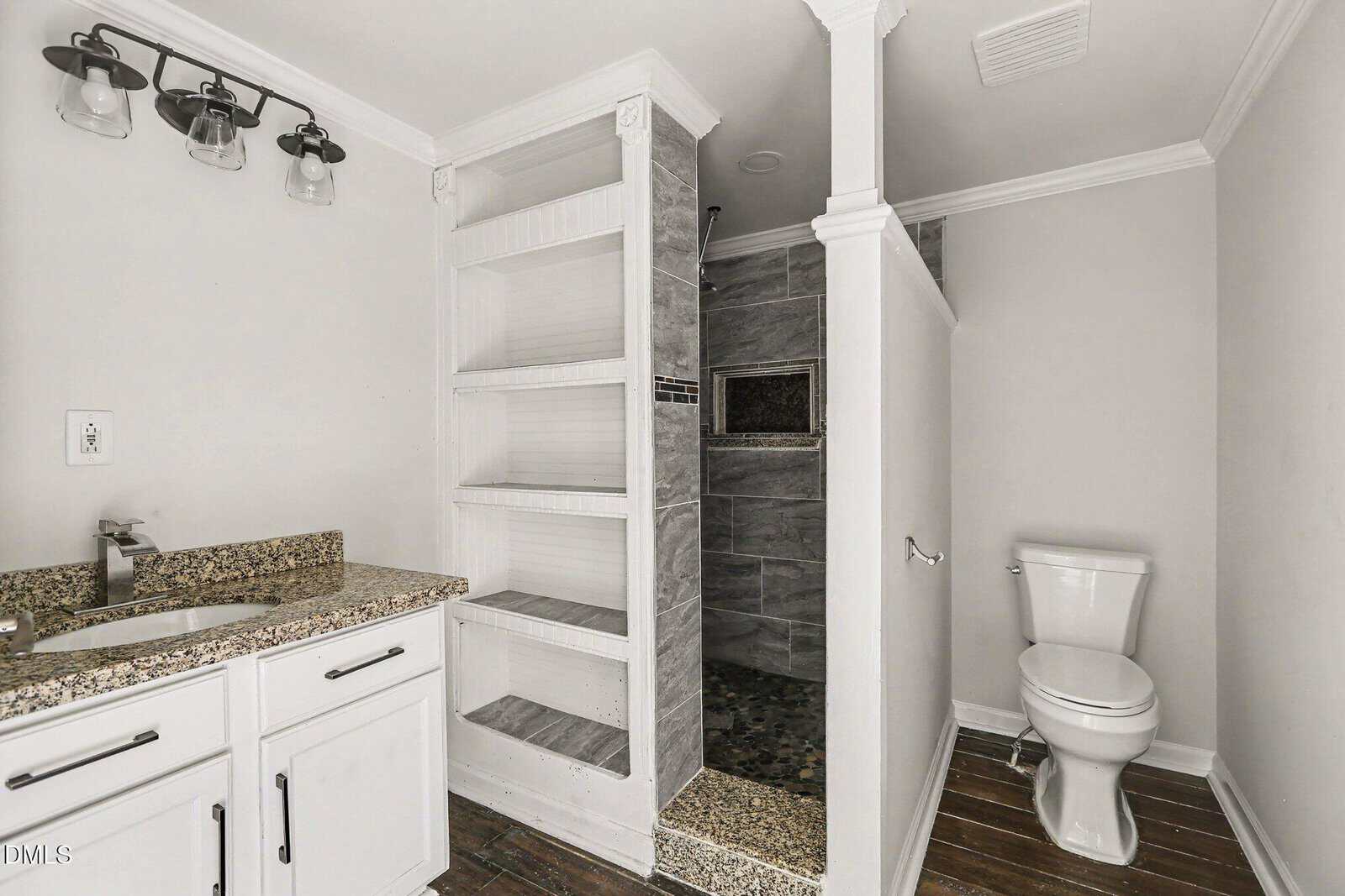 A modern bathroom with a white vanity and granite countertop, built-in open shelves, a walk-in gray tiled shower, and a white toilet on dark wood flooring.