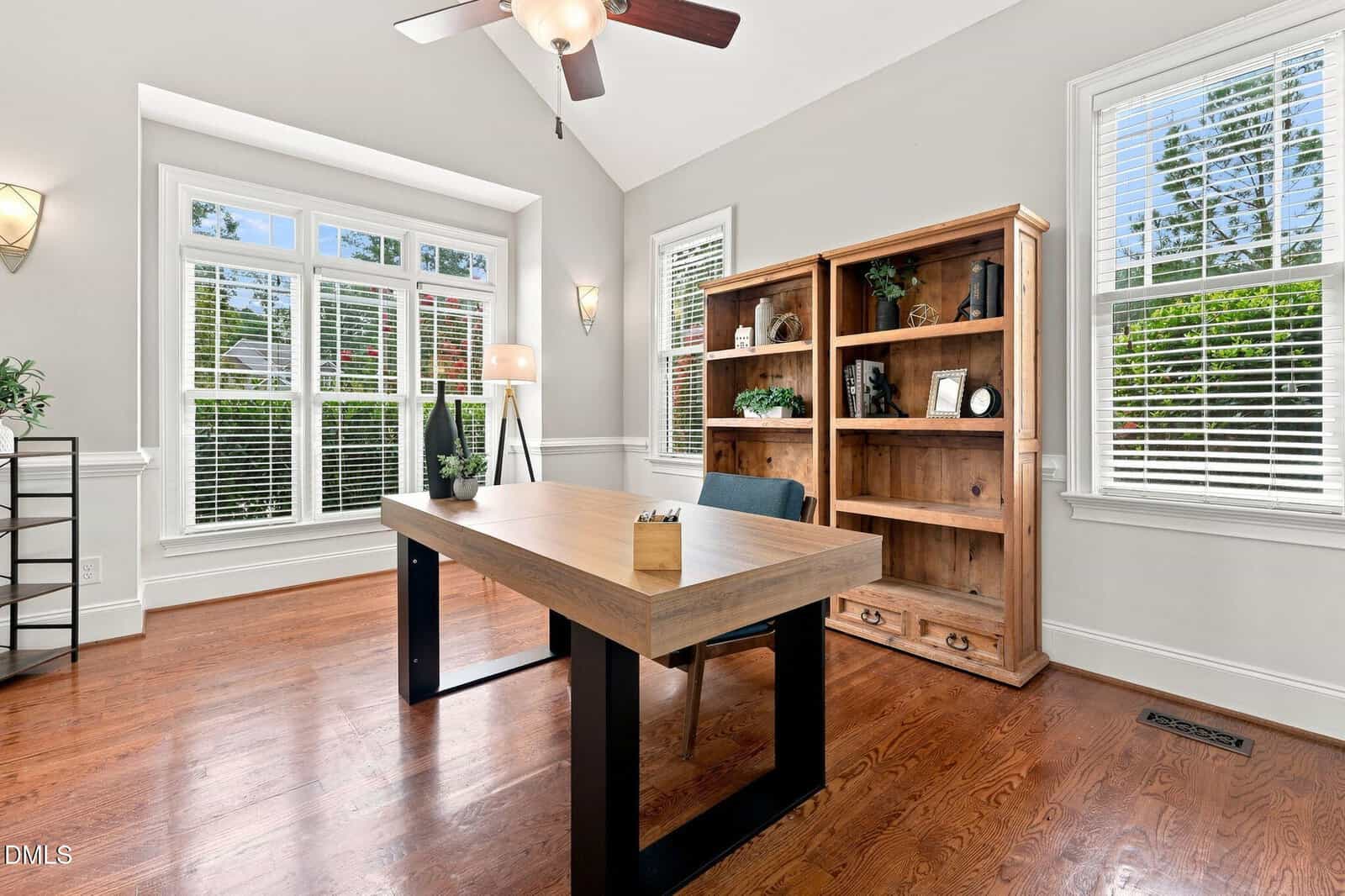 Home office with a wooden desk, black chair, large wooden bookshelf, floor lamp, and decorative items. White walls, hardwood floors, and windows with blinds let in natural light. Ceiling fan is visible above.