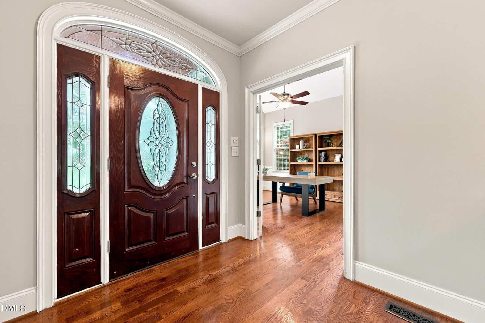 A wooden front door with decorative glass panels opens to a hallway with wood floors. An adjacent room has a ceiling fan, large window, bookshelf, and desk. Walls are light-colored and trim is white.