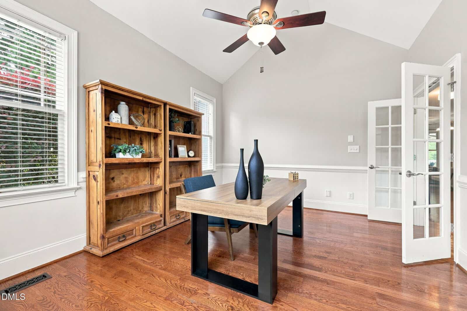 A bright office with wood floors, a wooden desk, two blue vases, a black chair, a wooden bookshelf with decor, a ceiling fan, and French doors. Light gray walls and white trim complete the room.