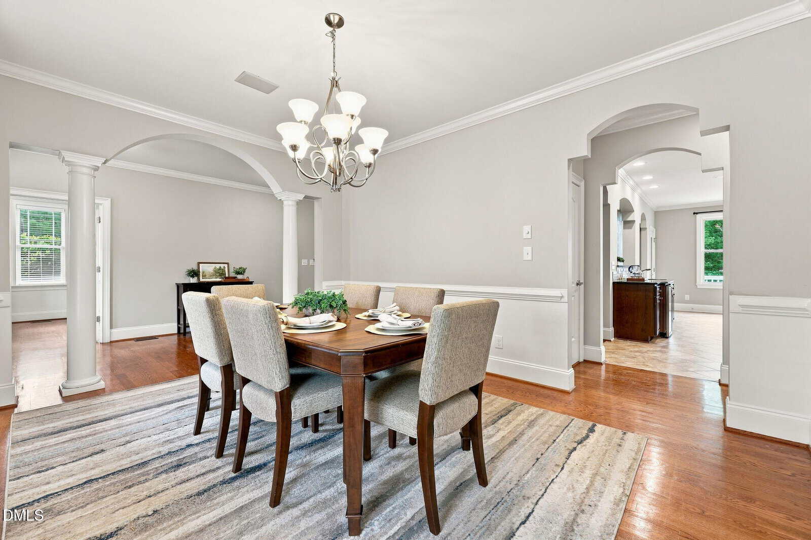 Bright dining room with a wooden table set for six, upholstered chairs, chandelier, striped rug, hardwood floors, arched doorways, and neutral walls. Large windows and columns add elegance to the space.