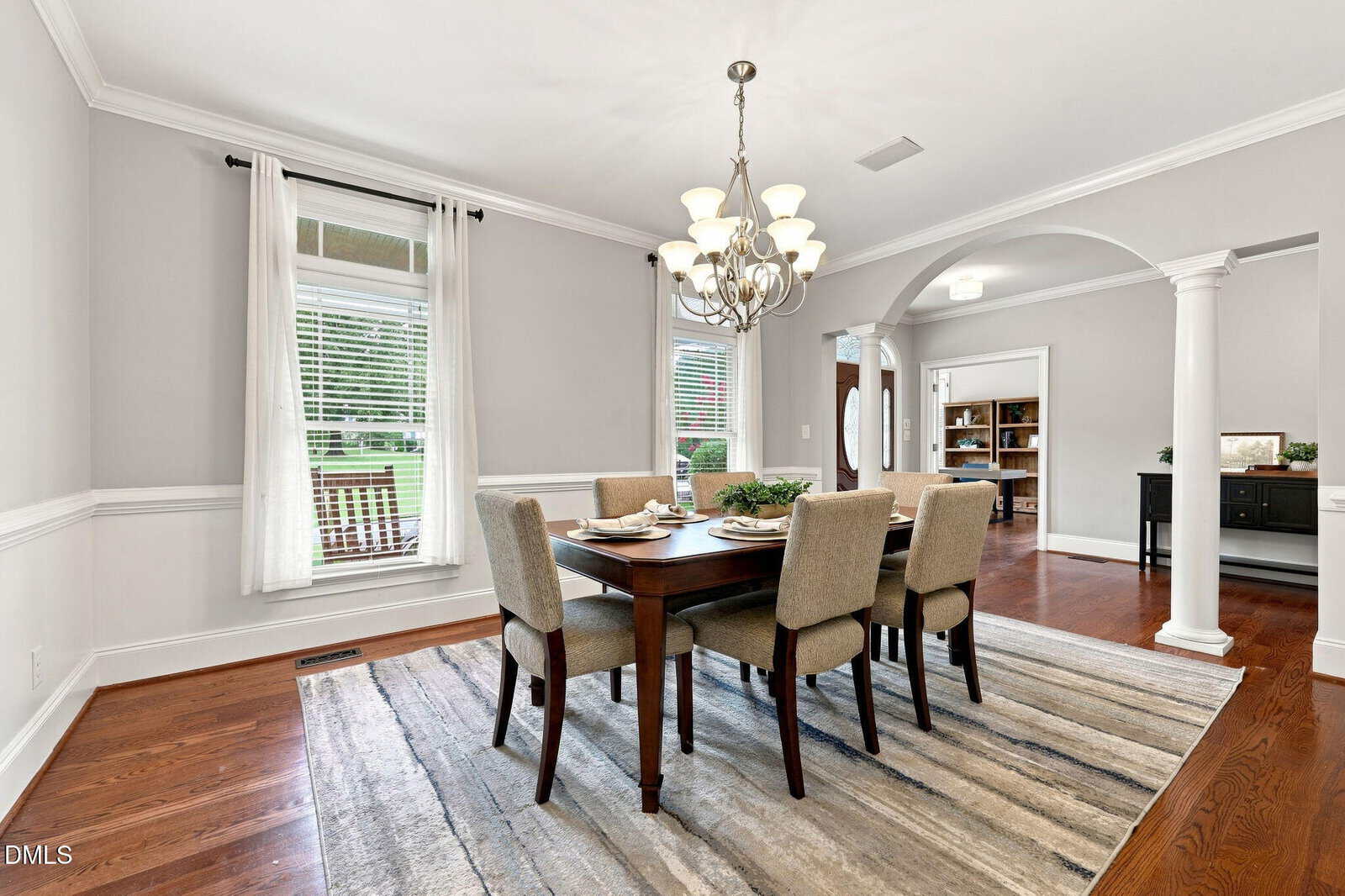 Elegant dining room with a wooden table set for six, beige upholstered chairs, a modern chandelier, hardwood floors, large windows with white curtains, and an archway leading to another room.