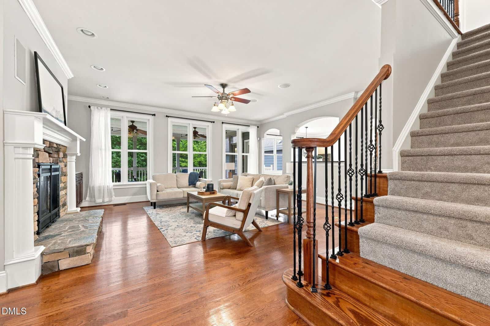 Bright living room with hardwood floors, stone fireplace, beige sofas, and large windows. Carpeted stairs with a wooden handrail are on the right. Ceiling fan and neutral decor give a cozy, welcoming feel.