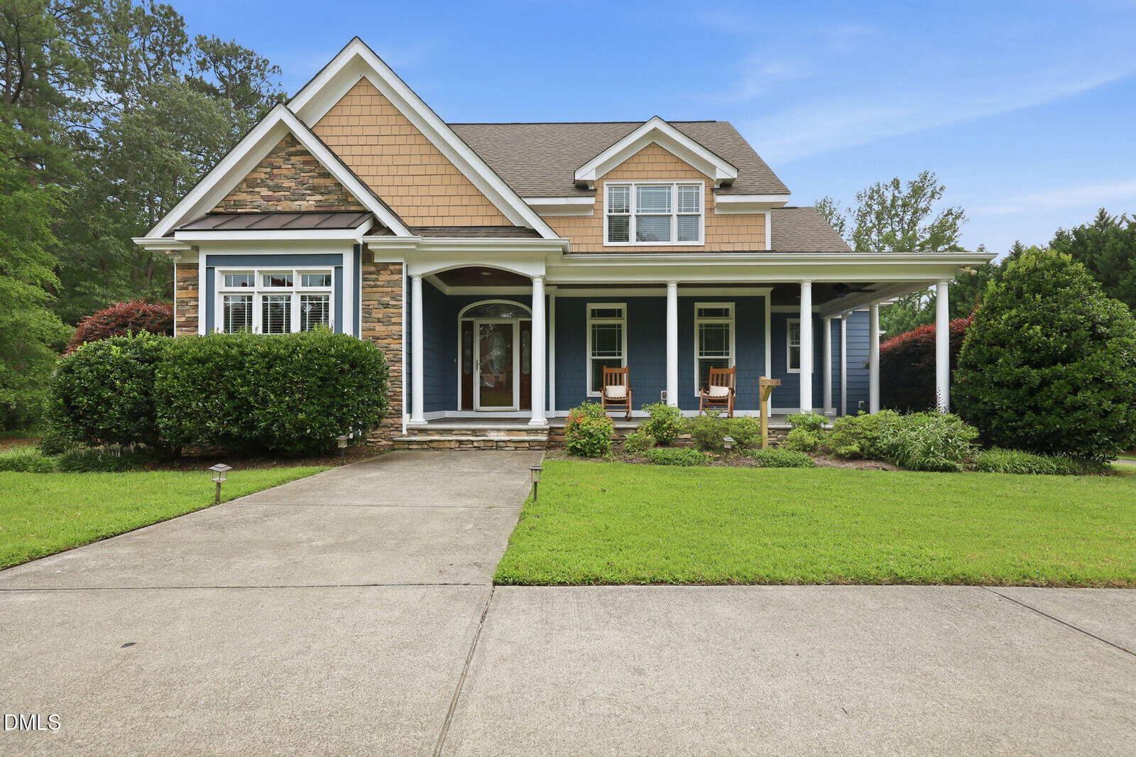 A two-story suburban house at 1530 Raven Wood Drive in Creedmoor featuring blue siding, tan shingle accents, stone detailing, and a front porch with white columns. A concrete driveway leads to the front entrance, which is flanked by green landscaping and two rocking chairs.
