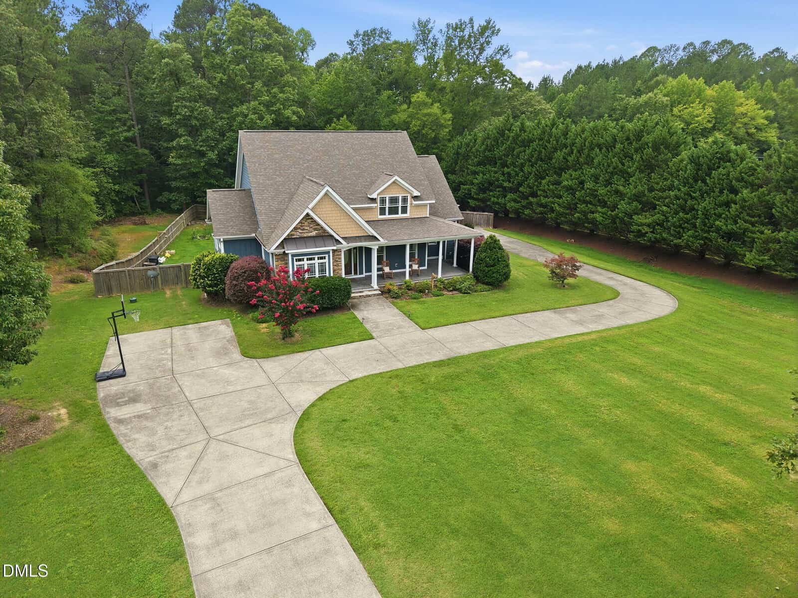 An aerial view of a two-story blue house with a covered front porch, a curved concrete driveway, a basketball hoop, and a large green lawn surrounded by dense trees.