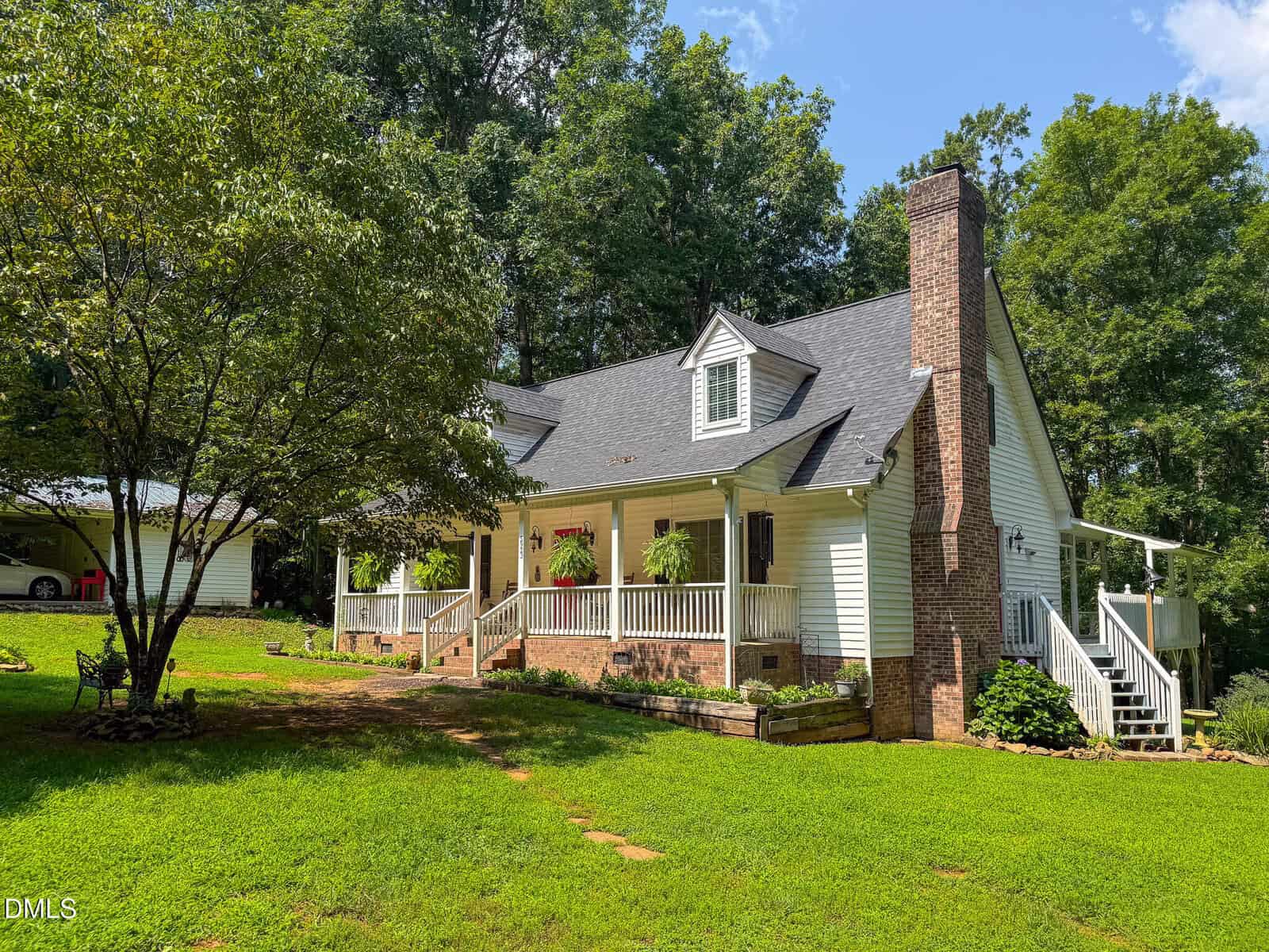 A charming white house with a brick chimney sits on a grassy lawn at 5923 Rivers Bend Drive, Rougemont, surrounded by trees. The house features a front porch with plants and steps, while a detached carport is visible in the background to the left.