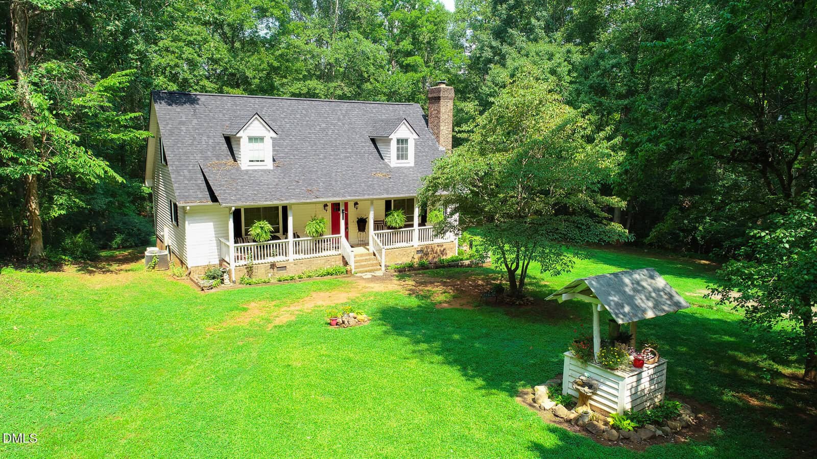 A white house with a covered front porch and dormer windows sits on a green lawn surrounded by trees. A small decorative well is in the yard, and flower pots decorate the porch.