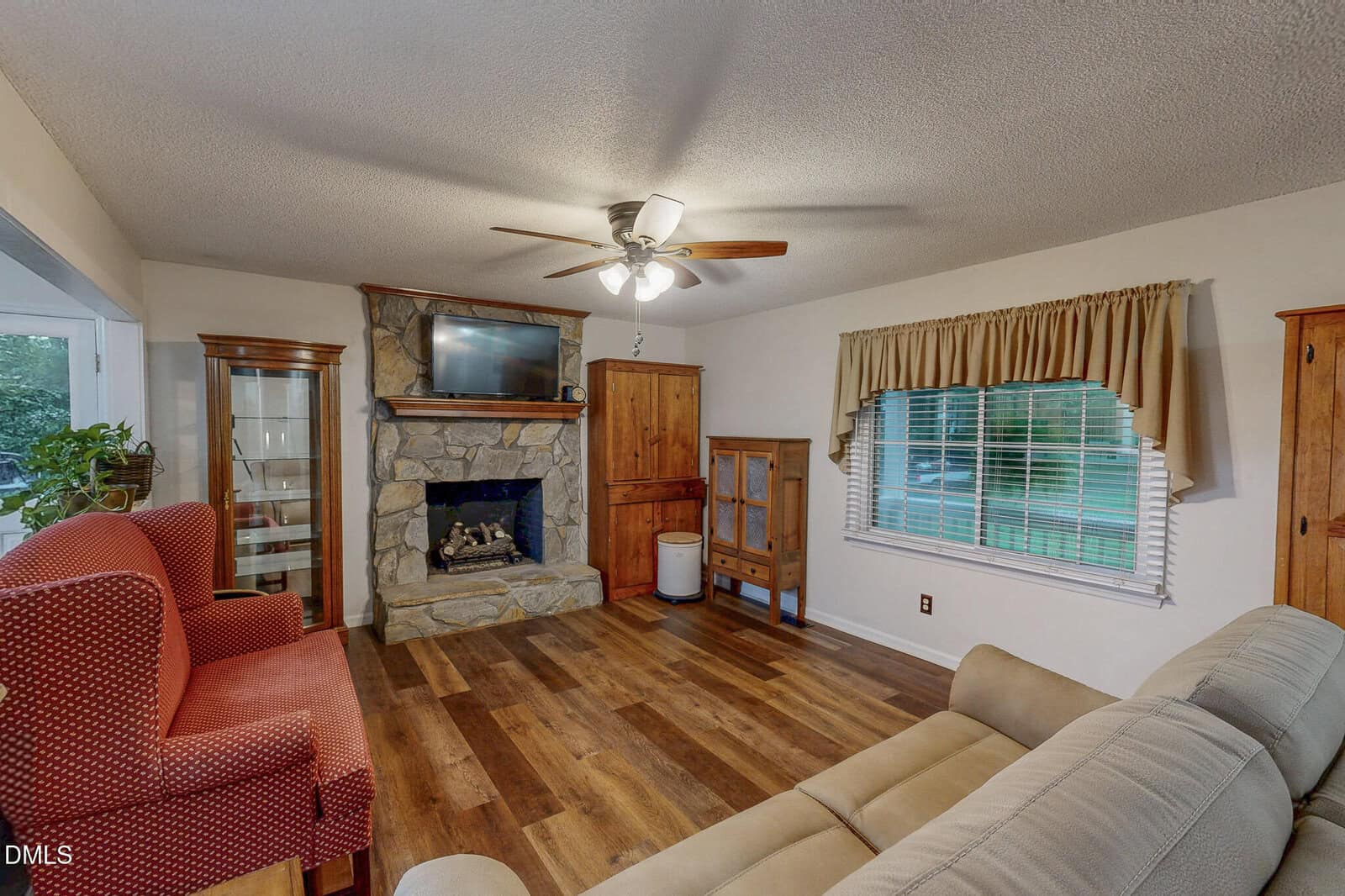 A living room with wood flooring, a stone fireplace, and a television mounted above the mantel. The room features a red patterned armchair, a light-colored sofa, a ceiling fan, and wooden cabinets. A window with a valance is on the right wall.