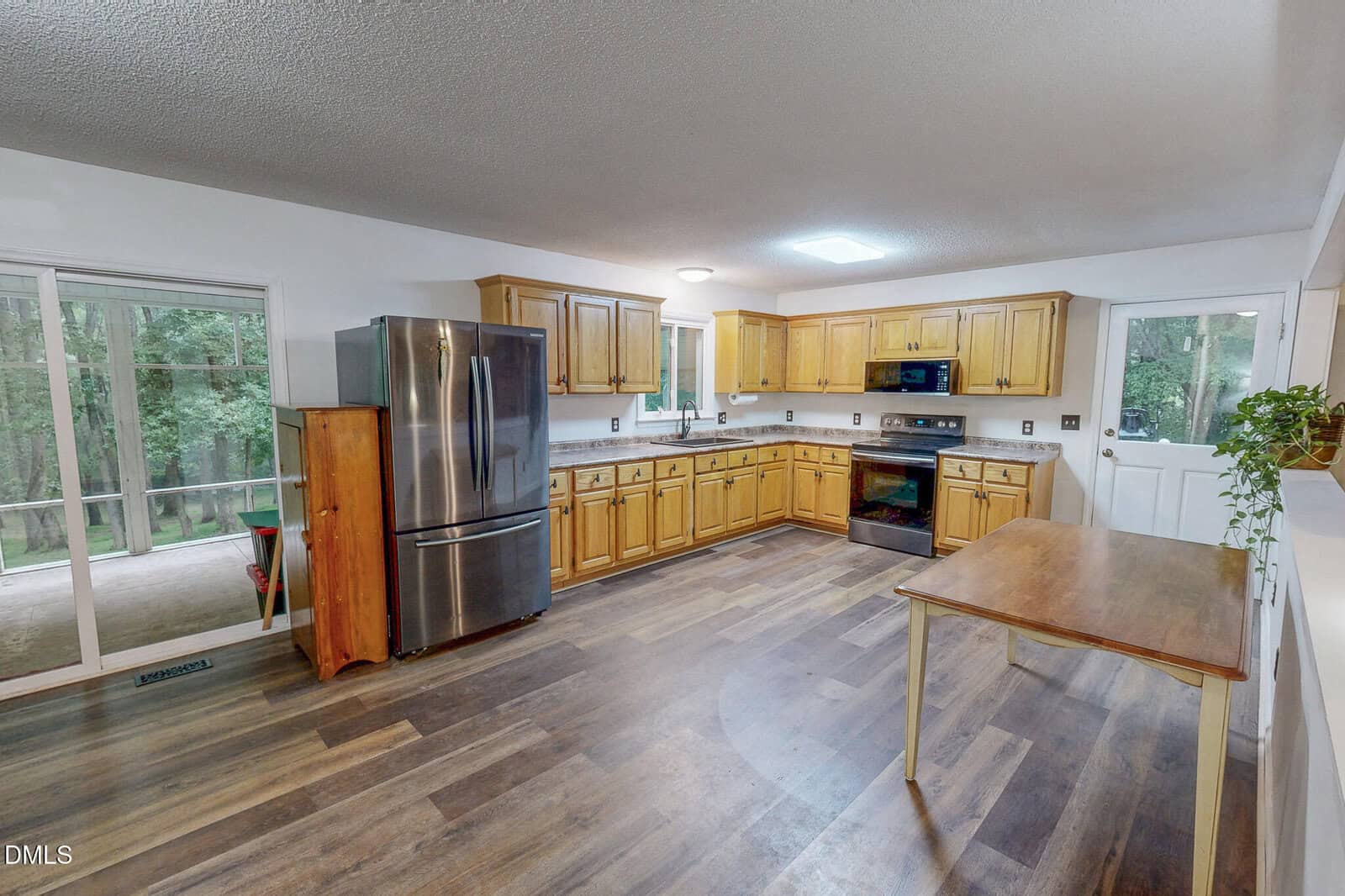 Spacious kitchen with wood cabinets, stainless steel refrigerator, electric stove, and dishwasher. Wood flooring, a wooden table, and a window over the sink. Glass door opens to a porch. White walls and ceiling.