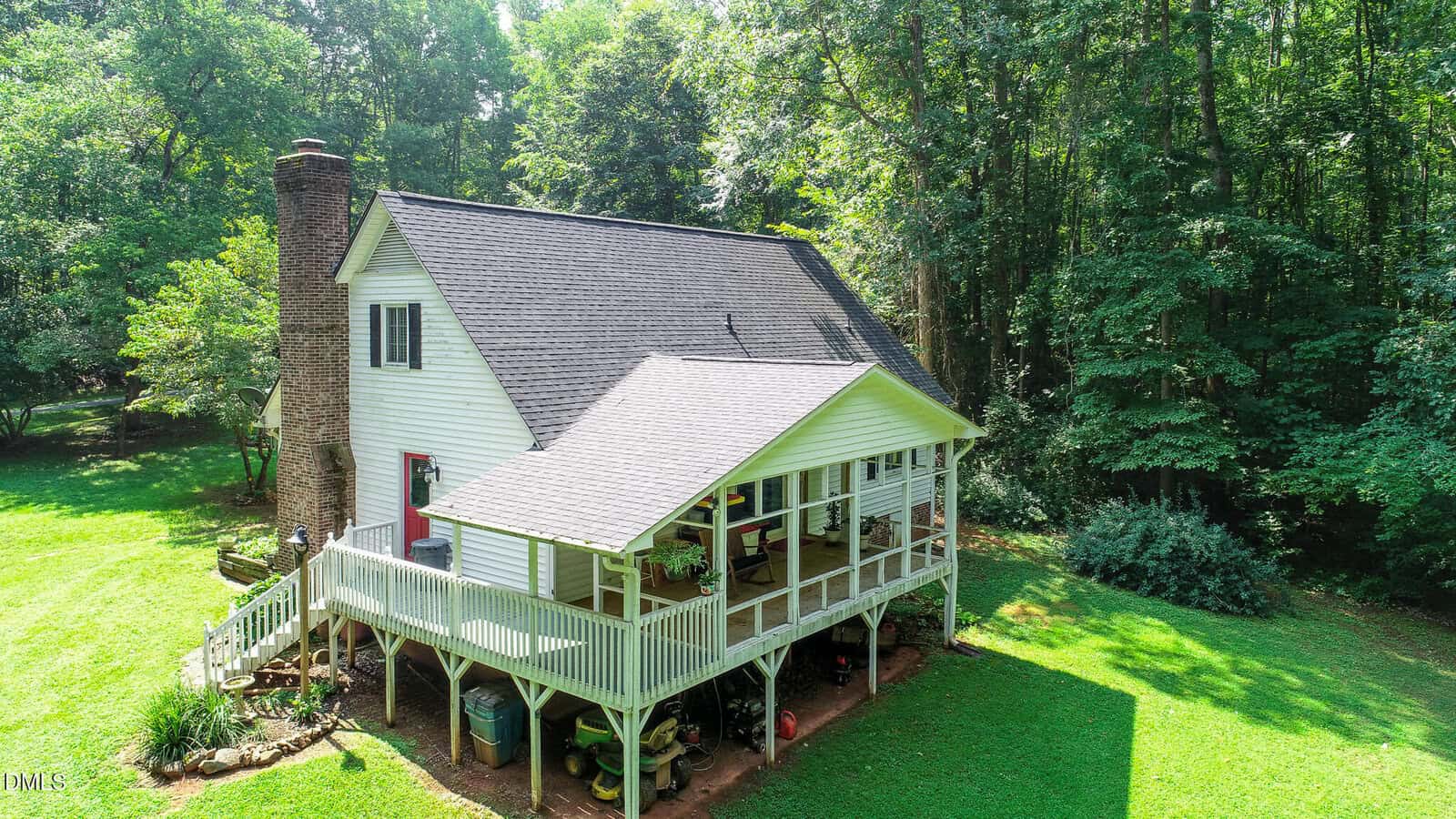 A white two-story house with a large wraparound porch sits on a grassy yard, surrounded by tall green trees, under bright sunlight.
