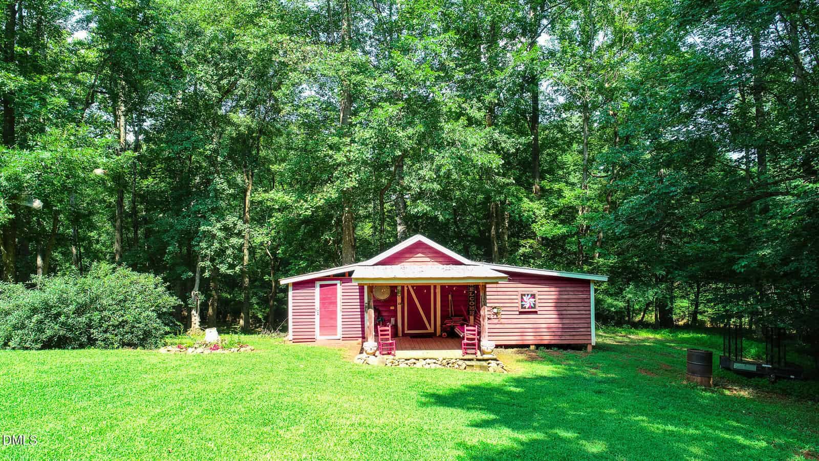 A small red cabin with white trim sits in a grassy yard, surrounded by tall trees. Two rocking chairs are on the front porch, and sunlight filters through the leaves, casting shade on the ground.