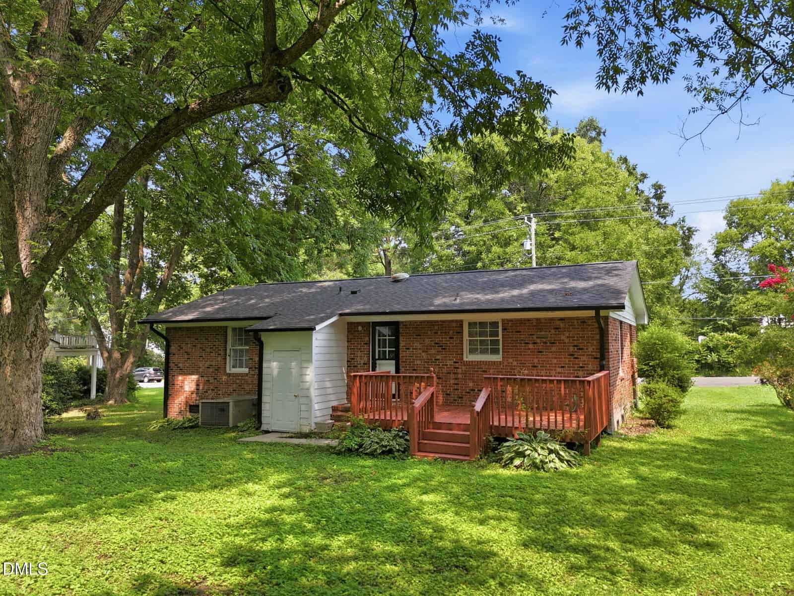 A small brick house with a gray roof, a wooden front porch with steps, surrounded by green grass and large trees under a clear blue sky.