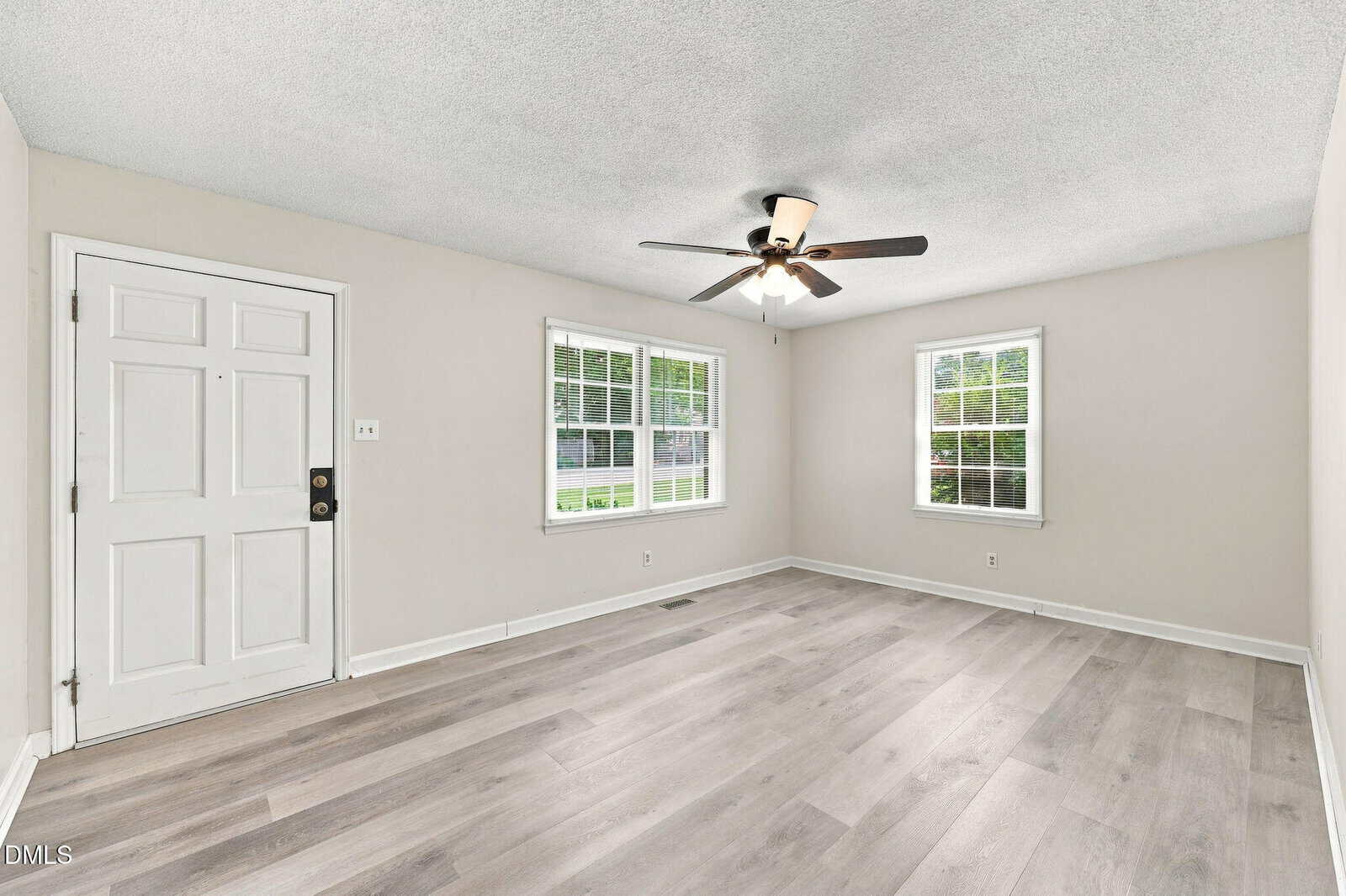 Unfurnished living room with light gray walls, wood-style flooring, two windows with white trim, a white front door, ceiling fan with lights, and natural light coming in. The room is empty and clean.