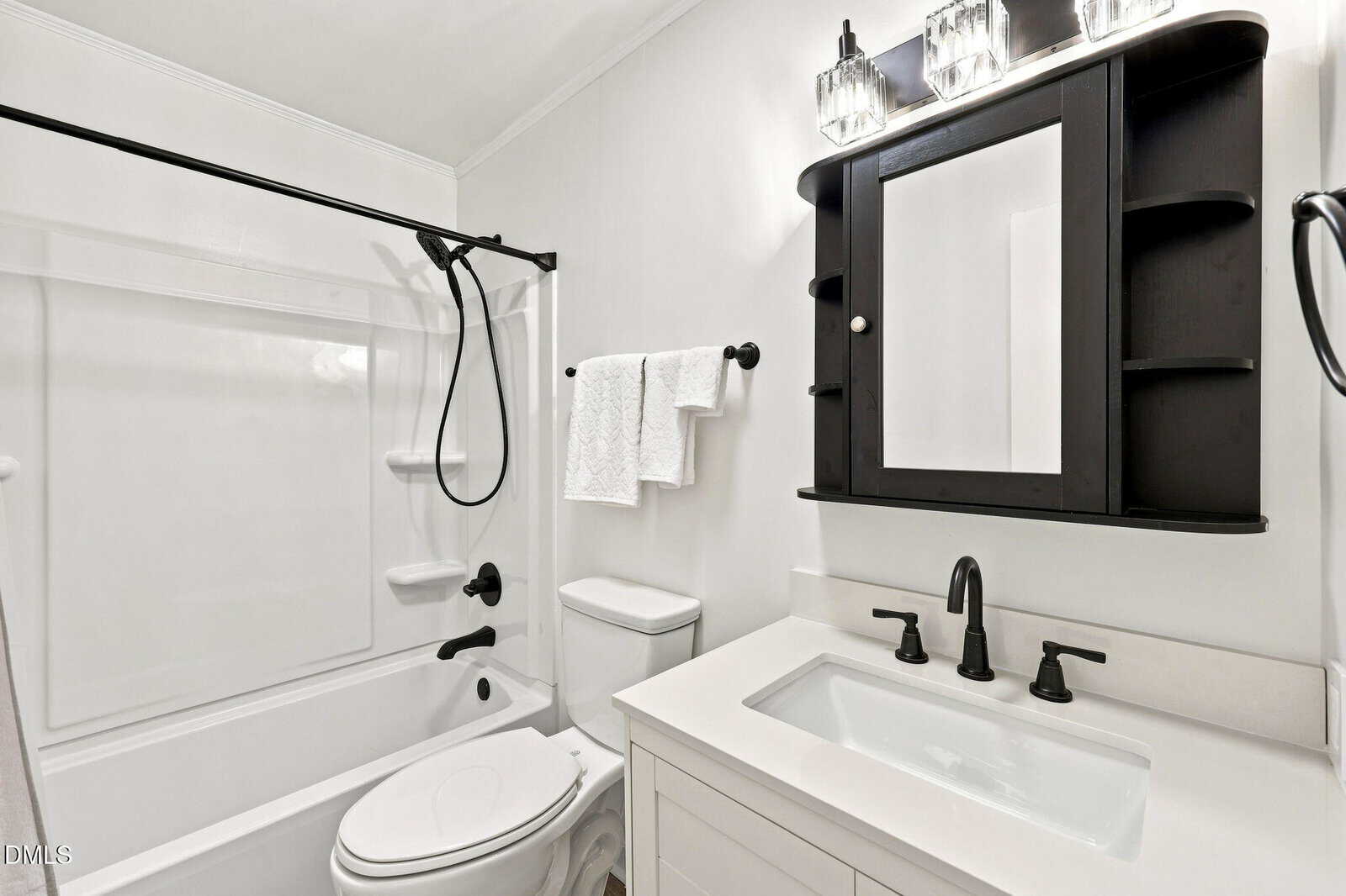 A modern bathroom with a white bathtub and shower, toilet, and white vanity. Black fixtures and a black medicine cabinet with lights above the sink contrast with the white walls and surfaces. Two towels hang on the rack.