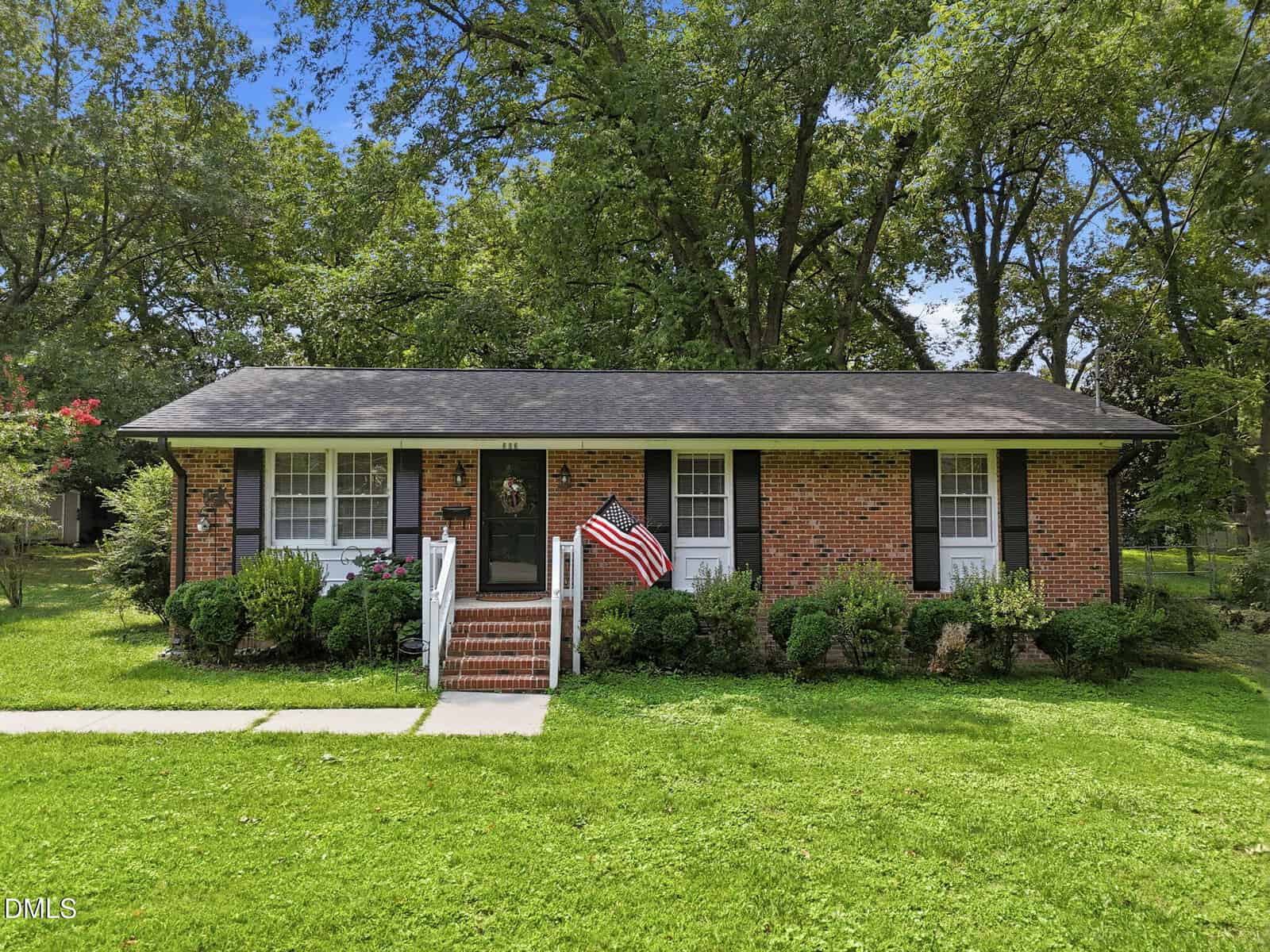 Single-story brick house with black shutters, a dark front door, steps leading to a small porch, an American flag, and green bushes in front, surrounded by a well-kept lawn and tall trees in the background.