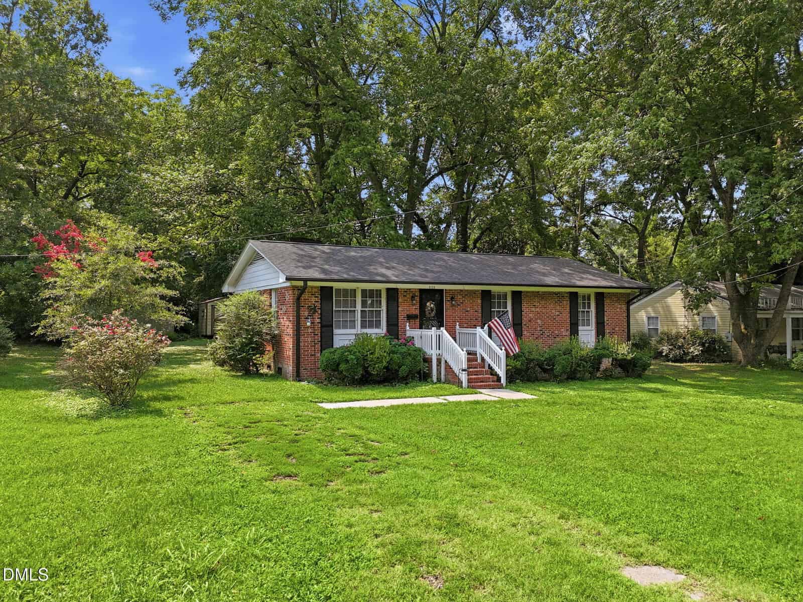 A small brick house with white trim sits on a well-kept lawn, surrounded by green trees and bushes, with a sidewalk leading to the front steps.
