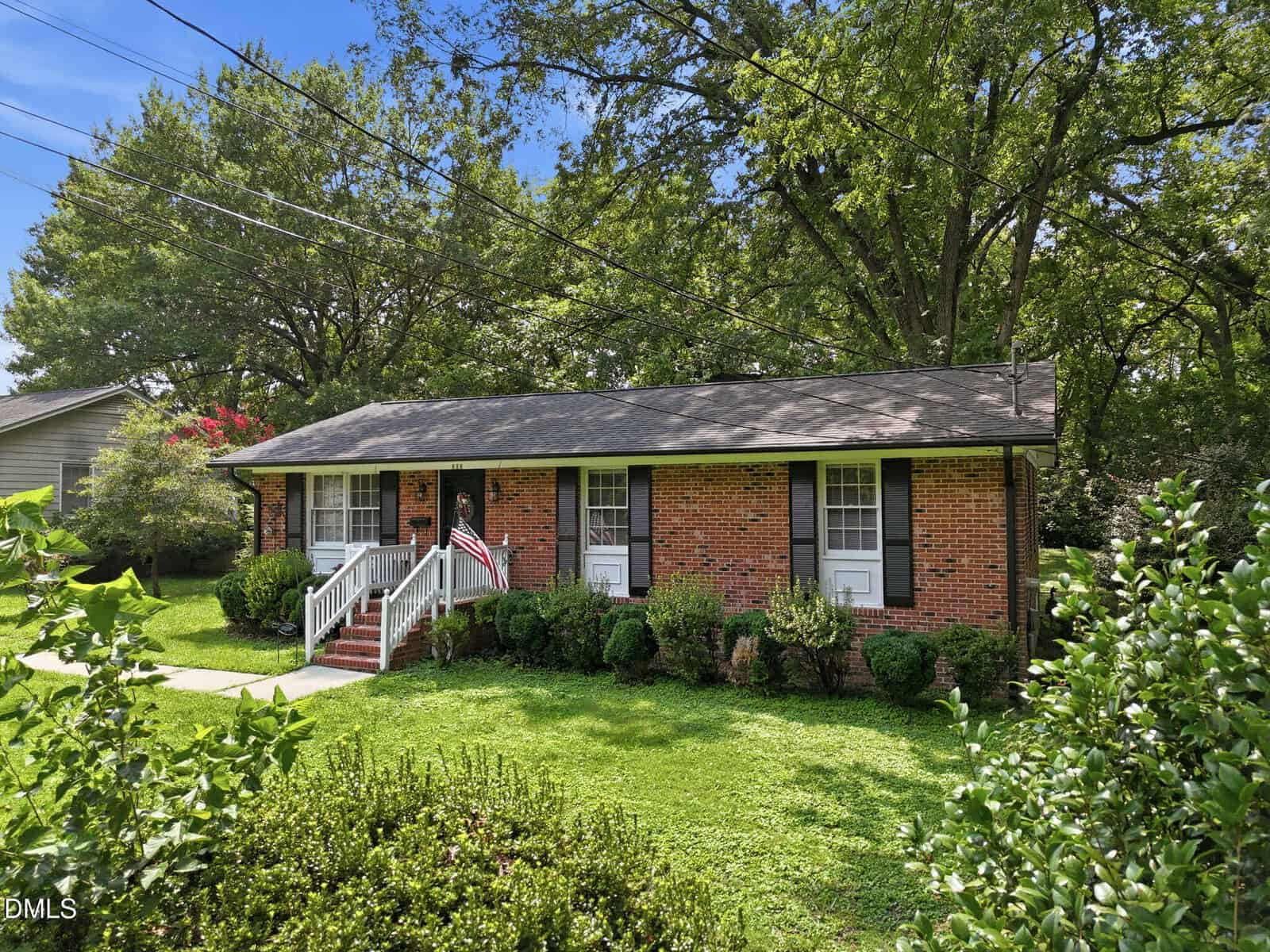 A one-story brick house with black shutters, white trim, and a front porch. Green shrubs and trees surround the house, and a well-kept lawn is in front.
