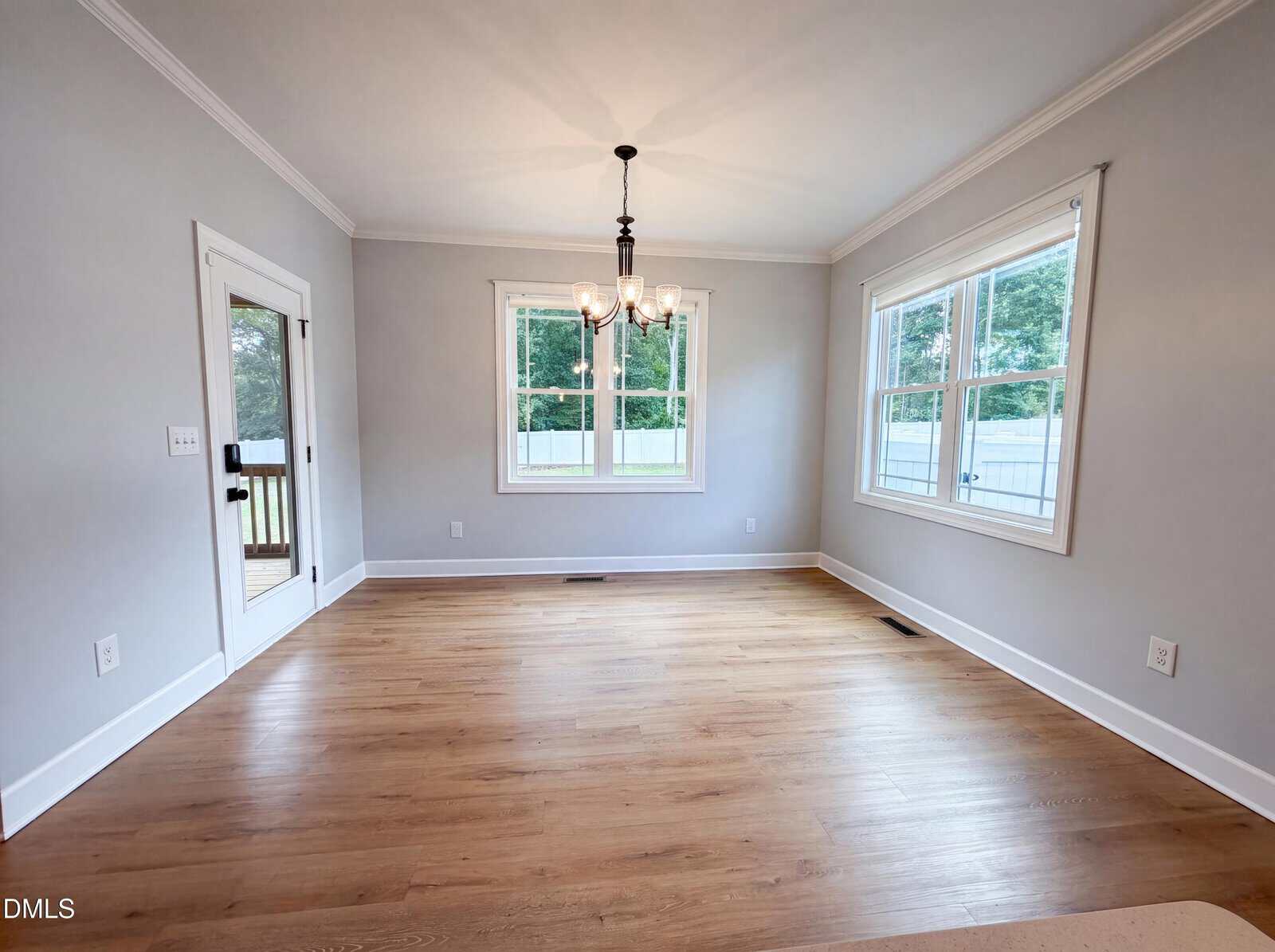 A bright, empty room with light gray walls, hardwood flooring, three large windows, a glass door to the outside, white trim, a black chandelier, and a white ceiling with crown molding.