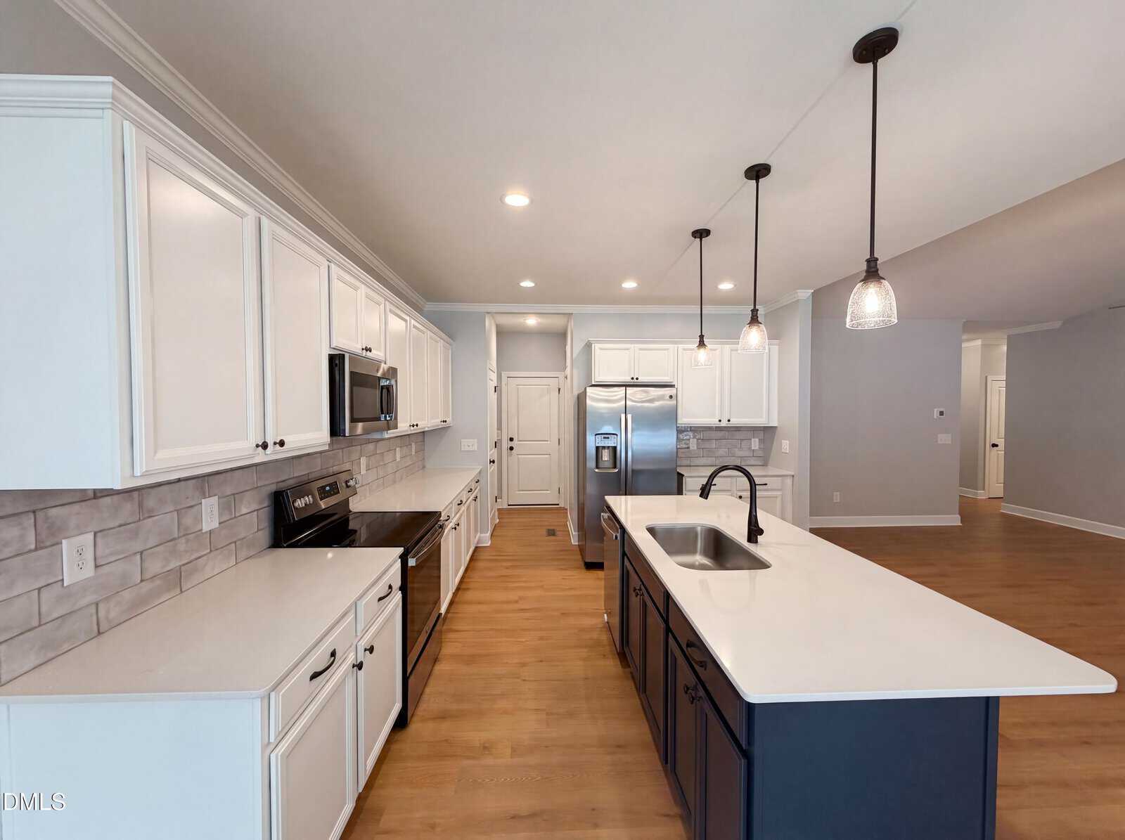 Modern kitchen with white cabinets, stainless steel appliances, a large center island with sink, pendant lights, tile backsplash, and wood flooring. Walls are light gray and overall space is bright and spacious.