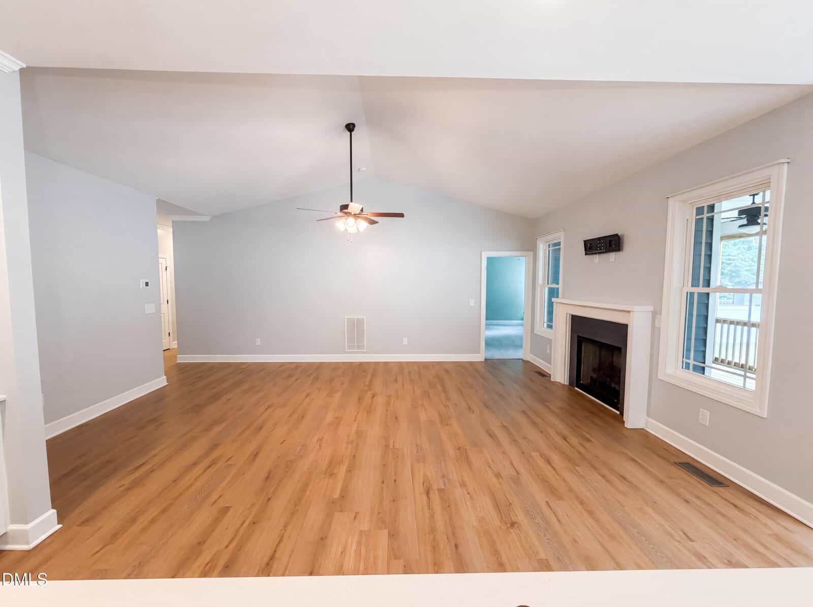 Spacious, empty living room with light gray walls, wood flooring, a ceiling fan with lights, a white fireplace, large windows, and a doorway leading to another room.