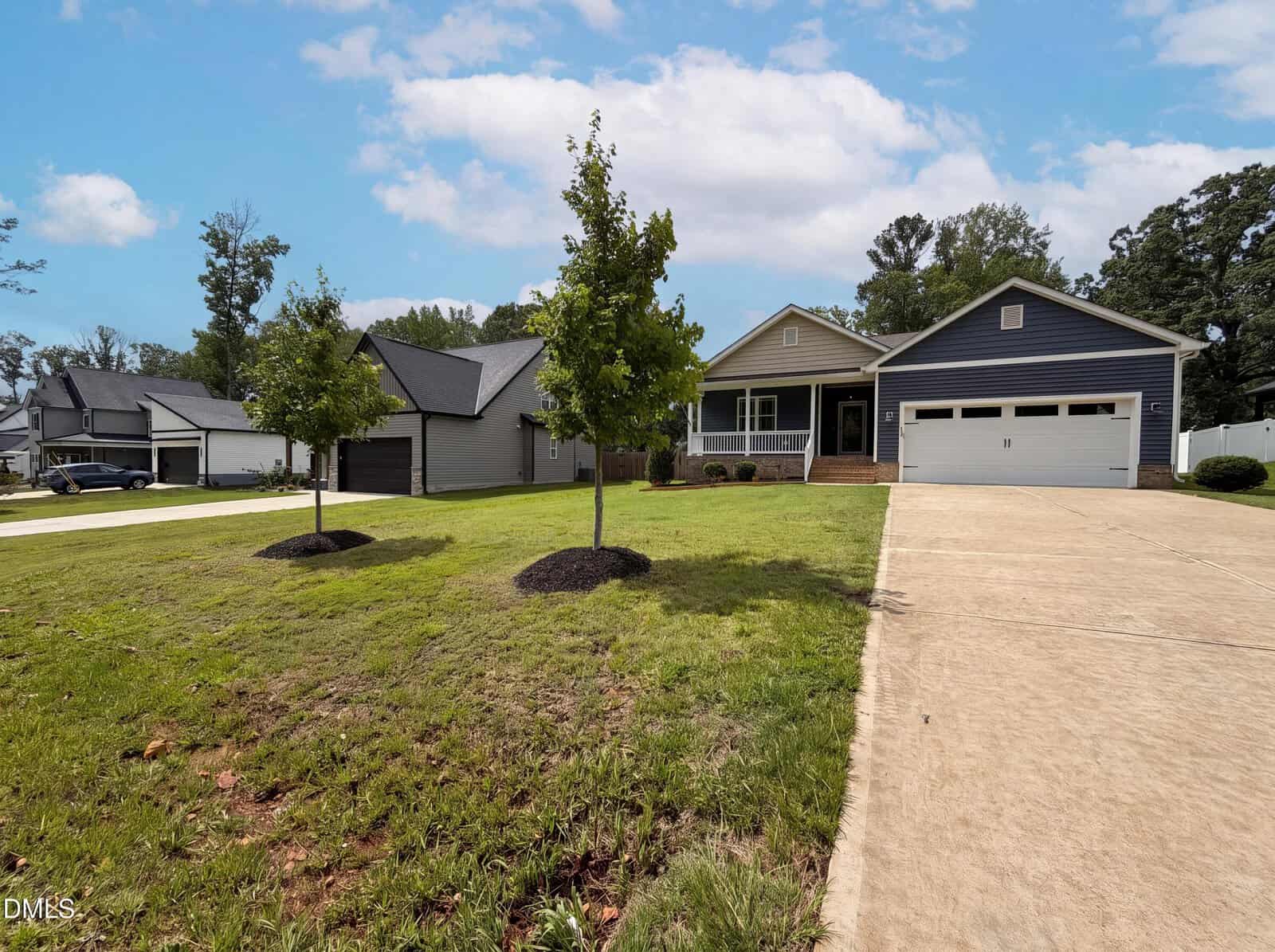 Single-story house with a blue and white exterior, attached two-car garage, front porch, concrete driveway, and a lawn with two young trees. Other houses are visible in the background under a partly cloudy sky.