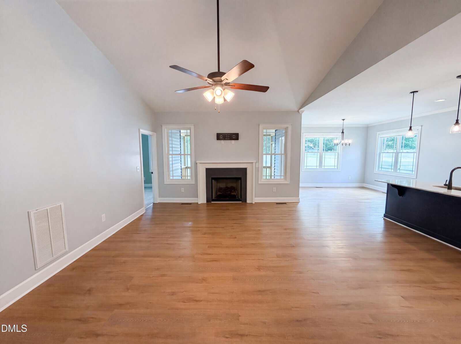 Spacious living area with light wood floors, ceiling fan, and fireplace centered on the far wall. Multiple windows provide natural light. Kitchen with island and sink is visible to the right. Walls are light gray.