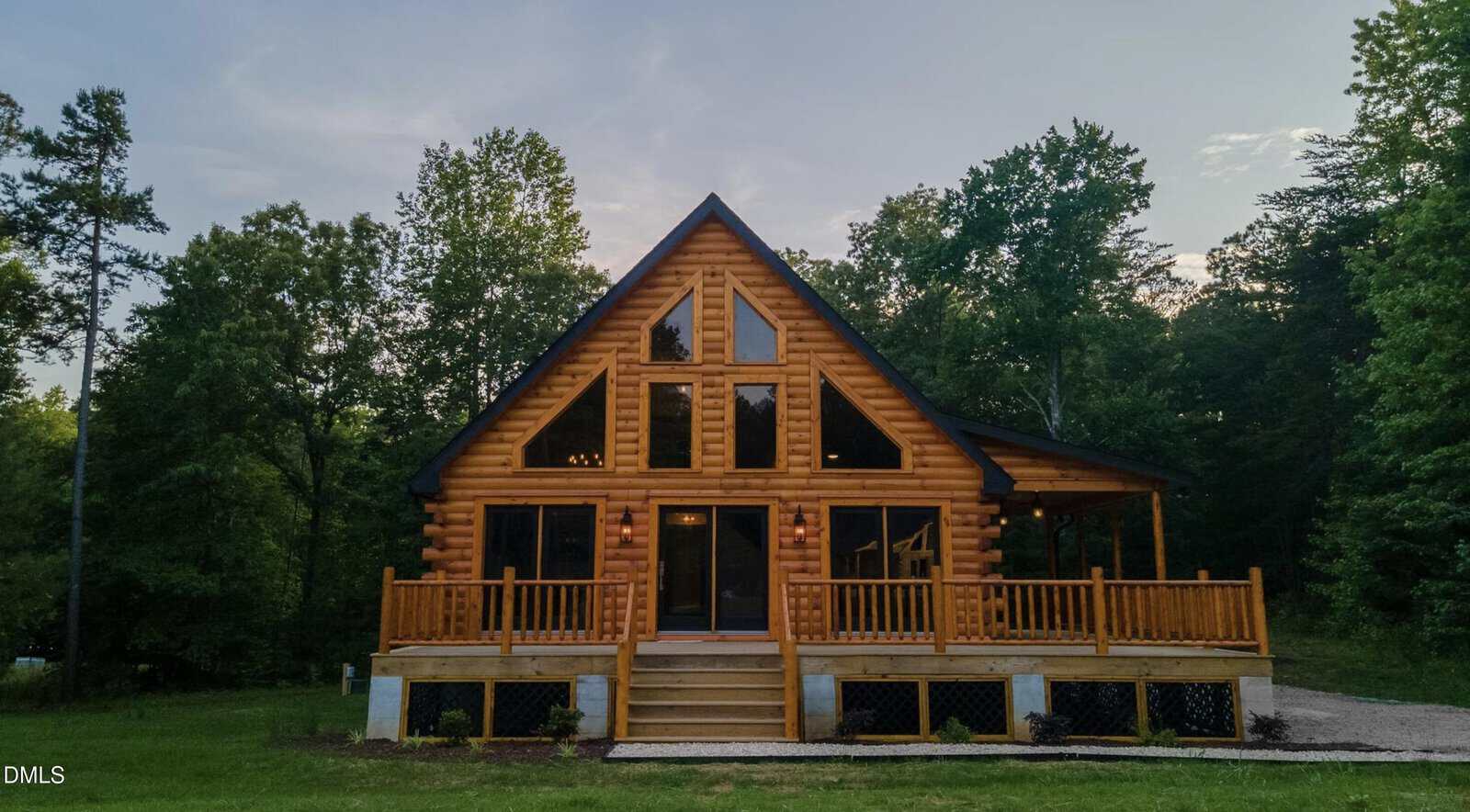 A log cabin with large triangular windows sits in front of a wooded area at 186 Buoy Drive, Henderson. The cabin has a front porch with railings, stairs leading to the entrance, and a peaked roof. The sky is clear with trees on both sides.