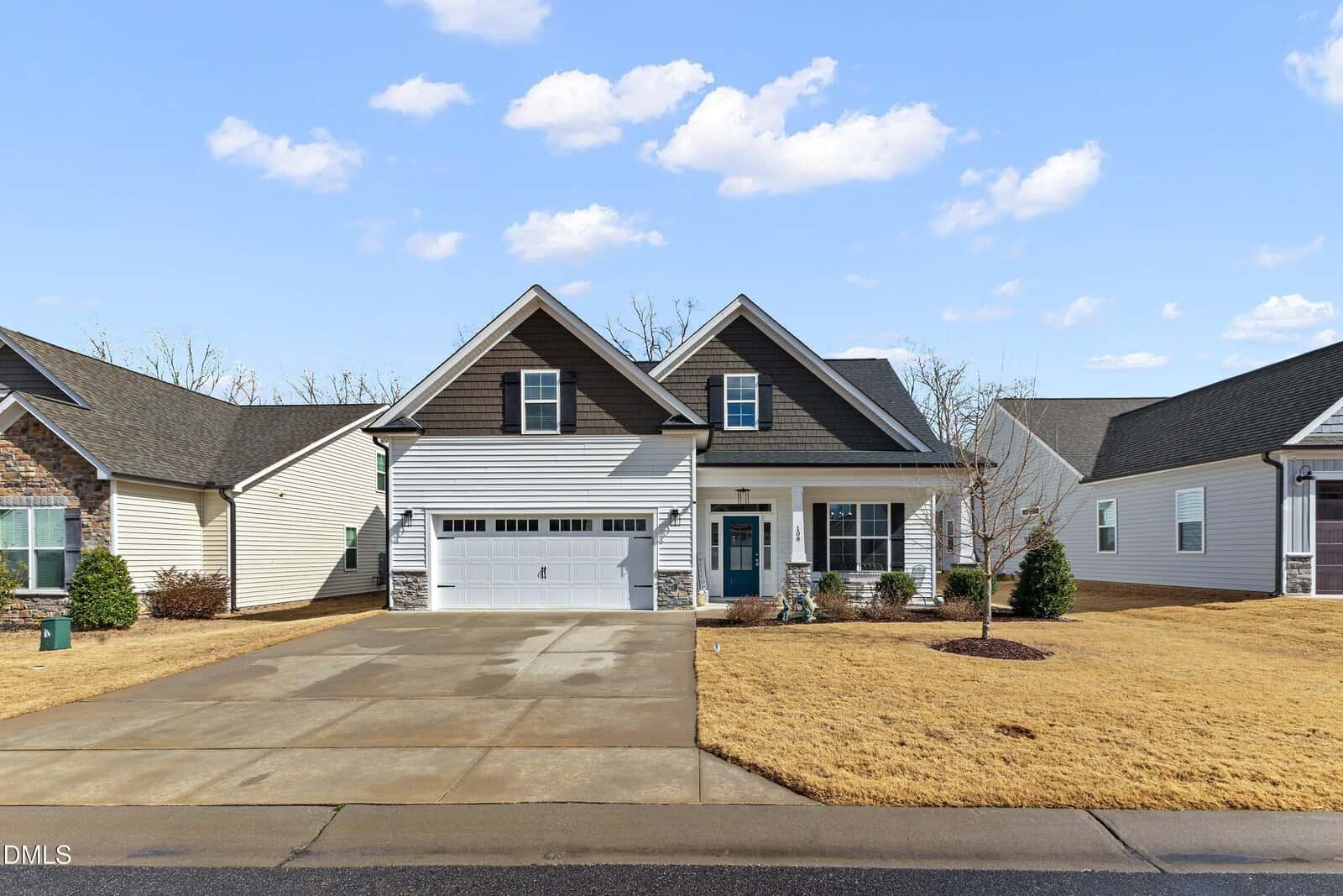 A modern, two-story suburban house with a front-facing garage, gray and white siding, gabled roof, and a wide driveway. The lawn is dry and yellowed, and the sky is partly cloudy.